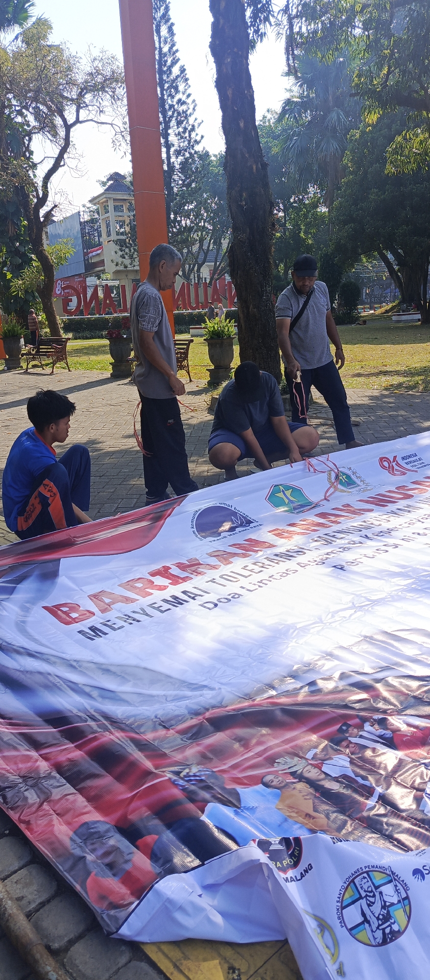 In a city park in Malang, Indonesia, four men are gathered around a large banner. The banner, which appears to be made of vinyl or a similar material, is spread across the paved ground. The men are in the process of handling the banner, with two of them appearing to adjust or fold it, while the other two observe.
The environment is outdoors, with trees and foliage visible in the background, suggesting a park setting. Sunlight illuminates the scene, casting shadows on the ground. There are buildings and structures in the distance, hinting at the urban landscape of Malang. The banner itself features large text in Indonesian, along with some logos and images. The text appears to be the main focus of the people's attention. The overall atmosphere is one of activity related to setting up or taking down the banner.