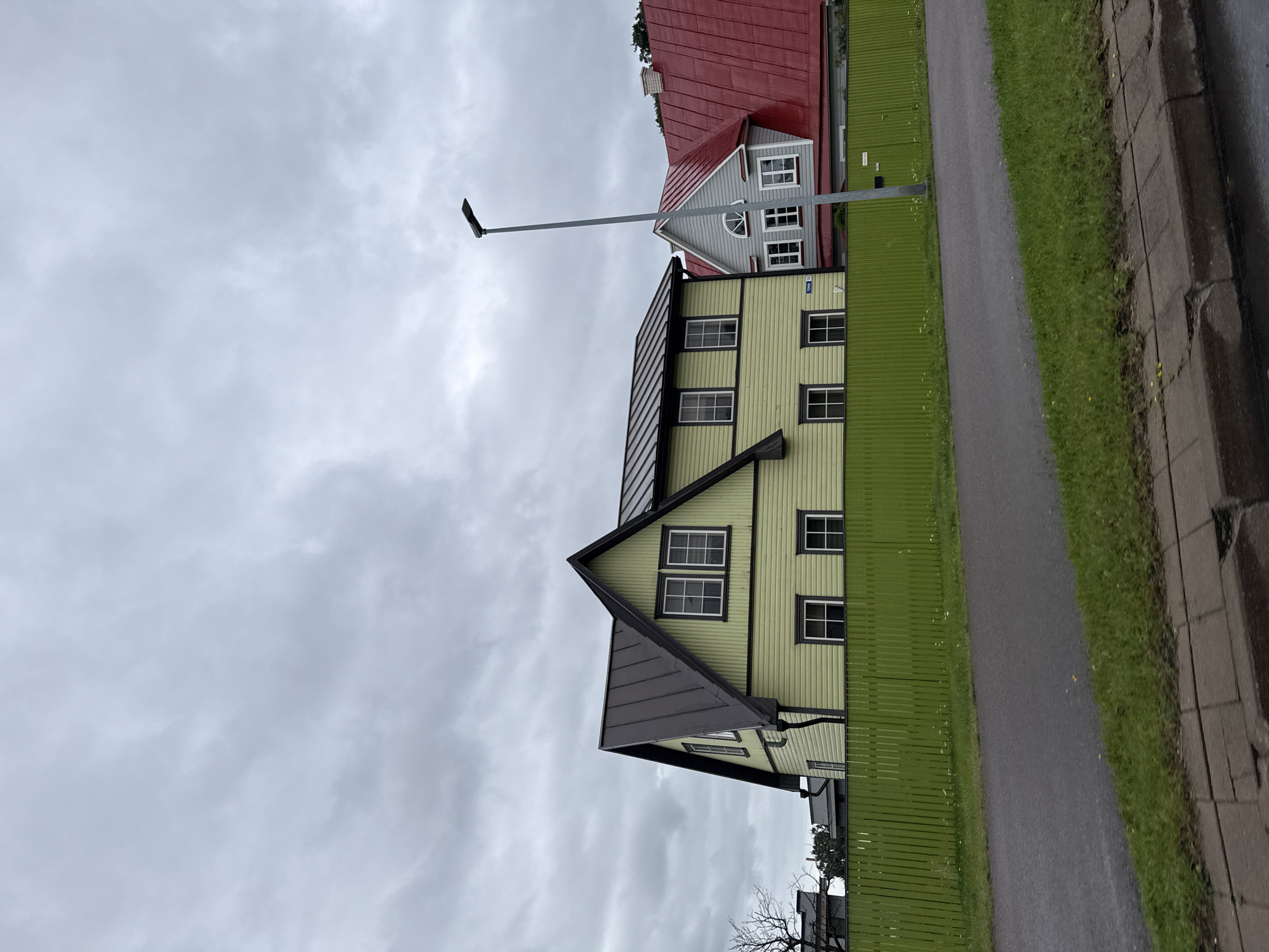 The image shows a street scene in Tallinn, Estonia, with two houses visible. The house in the foreground is a light green, two-story building with a dark metal roof and dark trim around the windows. It has multiple windows with white panes. A bright green wooden fence runs along the front of the property. To the right of the green house is another house with a bright red metal roof and gray siding. This house also has multiple windows. A tall, thin street lamp stands between the two houses. The sky is overcast with gray clouds. The foreground shows a paved road and a grassy area with a sidewalk. The overall impression is a residential street on a cloudy day.