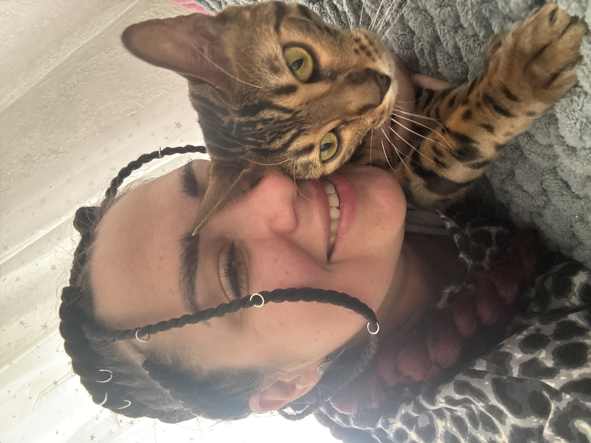 An overhead close-up shot shows a person with dark braided hair, some adorned with small silver hoops, embracing a Bengal cat. The person is smiling with their eyes closed, and their face is partially obscured by the cat, which is positioned on the person's shoulder and nuzzling their cheek. The cat has distinctive spotted fur and bright yellow-green eyes. The background is out of focus, with a patterned curtain or sheet visible. The person is wearing a leopard-print top. The overall impression is one of affection and closeness between the person and the cat.