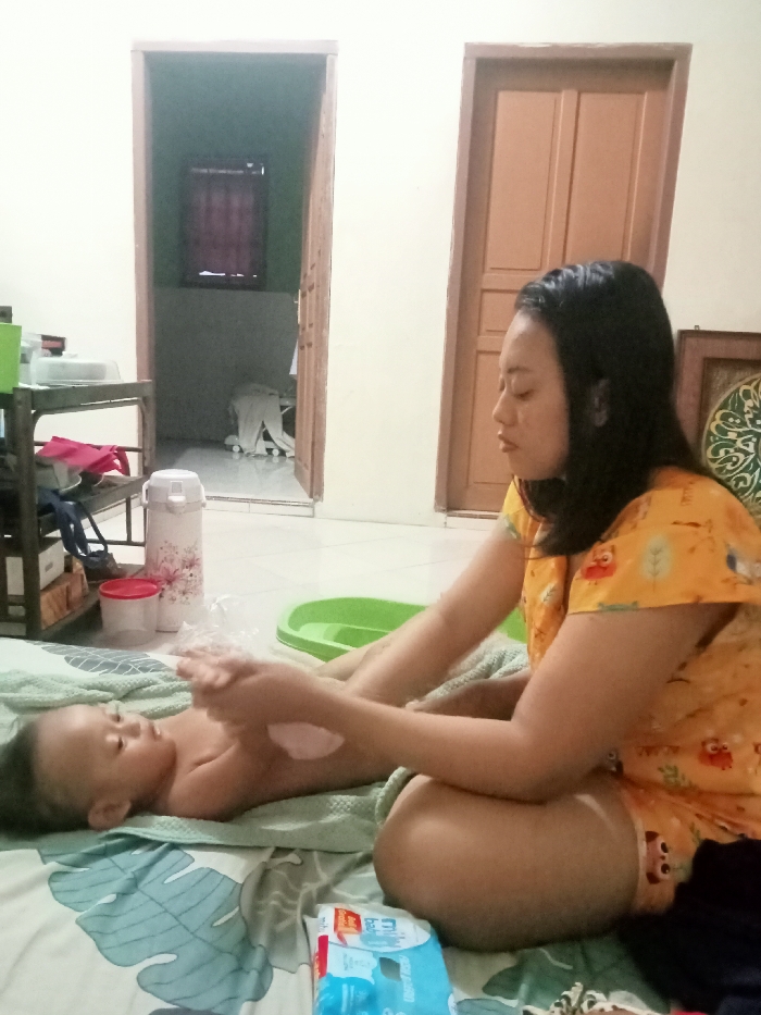 A woman applies lotion to a baby lying on a bed, both situated indoors. The woman, wearing an orange patterned top, is kneeling beside the child. Nearby, a green basin, water bottle, and other supplies are visible. In the background, an open doorway leads to another room, and a closed wooden door is present on the right. A small metal shelving unit with various items is located in the background to the left.