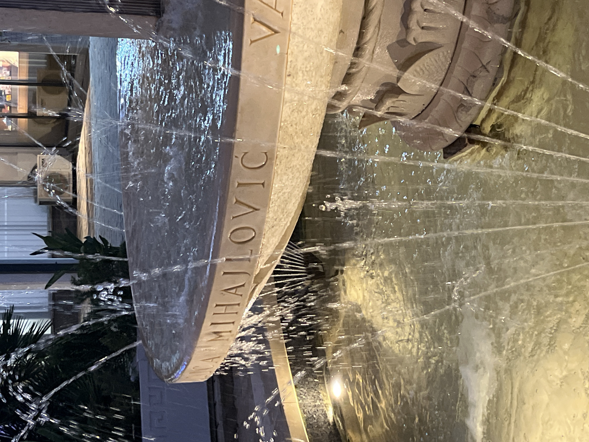 The image shows a close-up of a fountain's stone rim, with water cascading over its edge and into the pool below. Engraved on the rim is text that reads, "MIHAJLOVIC VA". The fountain is made of light-colored stone, with ornate carvings visible on the lower section. The water is illuminated, creating reflections in the pool. In the background, plants and building facades are visible, suggesting an outdoor urban setting. The lighting suggests it is evening or nighttime. The fountain is likely located in the city of Budva, Montenegro, as indicated by the name "MIHAJLOVIC," which is a common surname in the region, and "VA" may stand for "Velika plaza" (Big Beach), a prominent location in Budva.