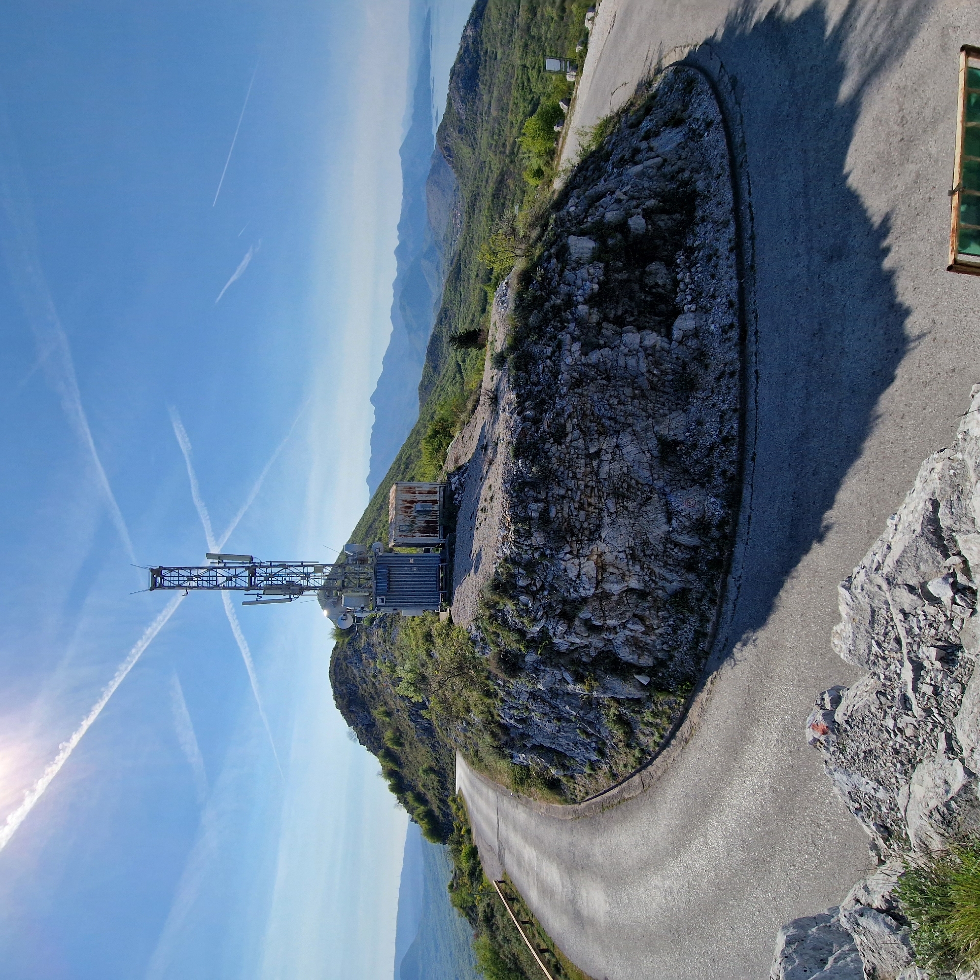 A low-angle, color photograph depicts a mountainous scene in Braiša, Brajsh, Montenegro. A communication tower is placed atop a rocky outcrop. The sky is light blue with multiple contrails. A winding road cuts around the outcrop, with a section of guardrail visible. The distant landscape features mountains and a body of water.
