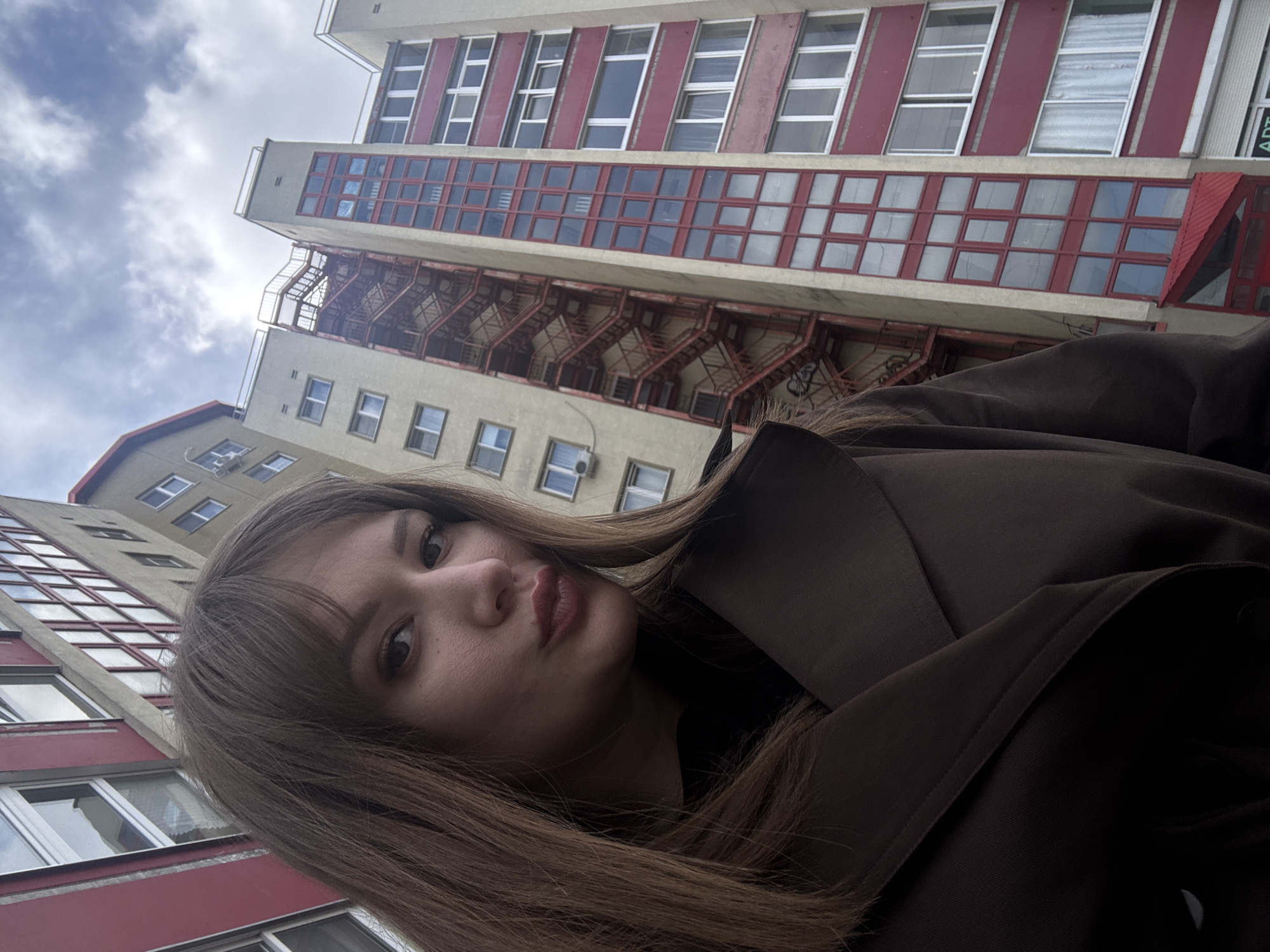 A selfie photo shows a woman with brown hair wearing a brown coat, positioned against a backdrop of buildings and sky. The buildings are multistory with windows and red frames, and one includes a fire escape. The sky is cloudy. The location is Chaikovsky, Russia.