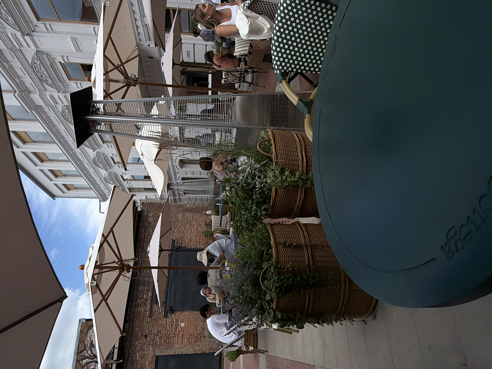 This media file captures an outdoor dining scene, likely a cafe or restaurant patio, with a distinctly European architectural backdrop. The image is oriented vertically. Several people are seated at tables, some engaged in conversation, others appearing to dine or relax.
In the foreground, a large, dark teal table dominates the right side of the frame, with a faint embossed logo visible on its surface. Adjacent to it are large woven baskets overflowing with greenery, suggesting a lush, natural element incorporated into the setting. Further back, white umbrellas provide shade for outdoor seating arrangements. People are seen scattered amongst these tables, with one woman in a white top and another in a light-colored top prominently featured.
The architecture in the background is detailed, with a classical style evident in the building's facade, featuring ornate carvings and multiple windows. The brickwork of an adjacent building adds a contrasting texture. The sky above is blue with some clouds, indicating it is daytime. The presence of a patio heater suggests it may be a cooler part of the day or a temperate climate.
The overall atmosphere appears relaxed and casual, consistent with an outdoor dining experience. The scene suggests a pleasant day for patrons to enjoy food and company al fresco. The crops offer closer views of the individuals, the table, and the building details, confirming the outdoor seating and the architectural elements.