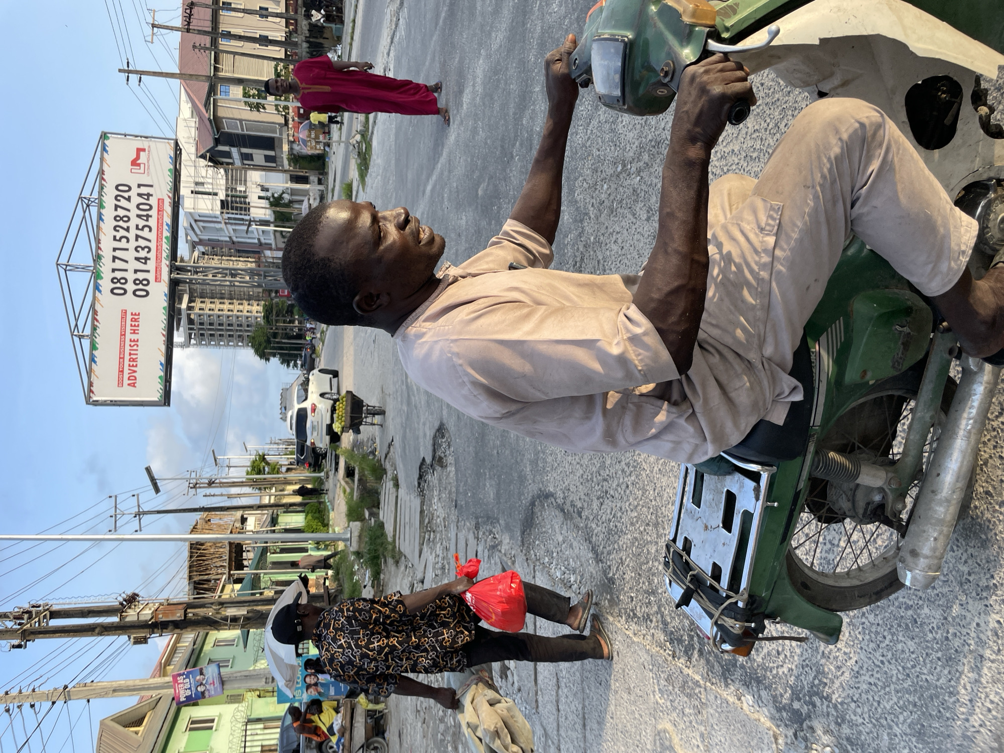 In Ilado, Nigeria, a man rides a green motorcycle on a paved street. He is wearing a light brown, loose-fitting outfit. The man is looking to his right, and his right hand is on the motorcycle's handlebars. Behind him, the motorcycle has a metal rack on the back. The street has cracks and patches.
To the left of the motorcyclist, another man walks away from the camera, carrying a large rolled-up bundle in his left arm and a red plastic bag in his right hand. He is wearing a dark patterned shirt and dark pants.
In the background, there is a white SUV on the right side of the street, and further down the street, a person in a red robe walks towards the camera. A billboard is visible above the buildings. The environment appears to be a bustling urban area with a mix of residential and commercial buildings, utility poles, and overhead wires. The sky is blue with some clouds.