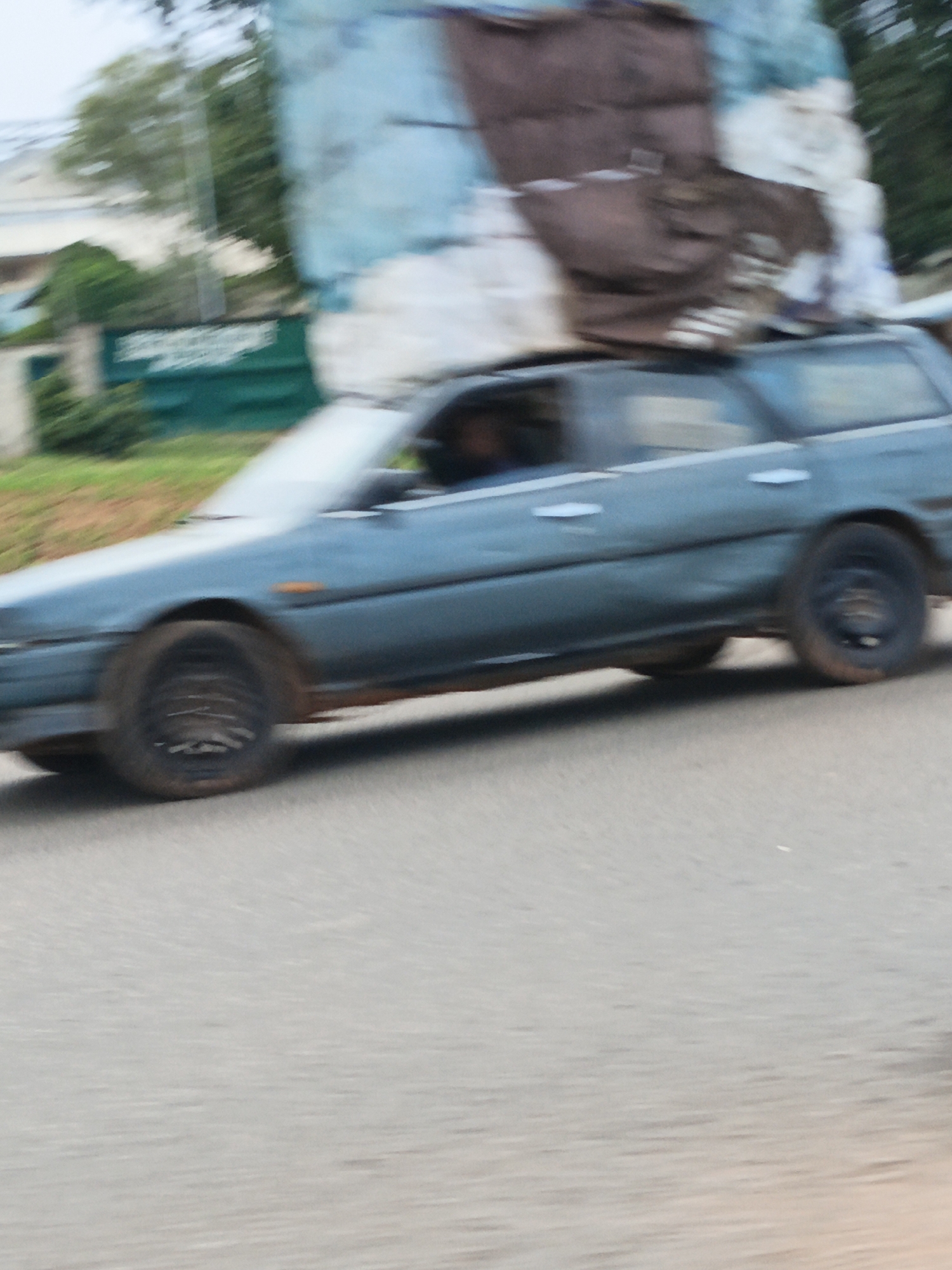 The image, captured in Kuru Dungus, Nigeria, shows a slightly blurred view of a dark blue station wagon driving on a paved road. The car is heavily laden with a large, bulky load secured on its roof, covered with a brown and light blue tarp. The load appears to be irregular in shape and texture. The car's windows are visible, with a dark figure, possibly a person, seen in the driver's seat. The car's wheels are dark, and the front passenger-side wheel is particularly visible. The background is indistinct due to motion blur but suggests an outdoor environment with greenery and buildings. The overall impression is of a vehicle in transit, likely carrying goods or belongings through the town.