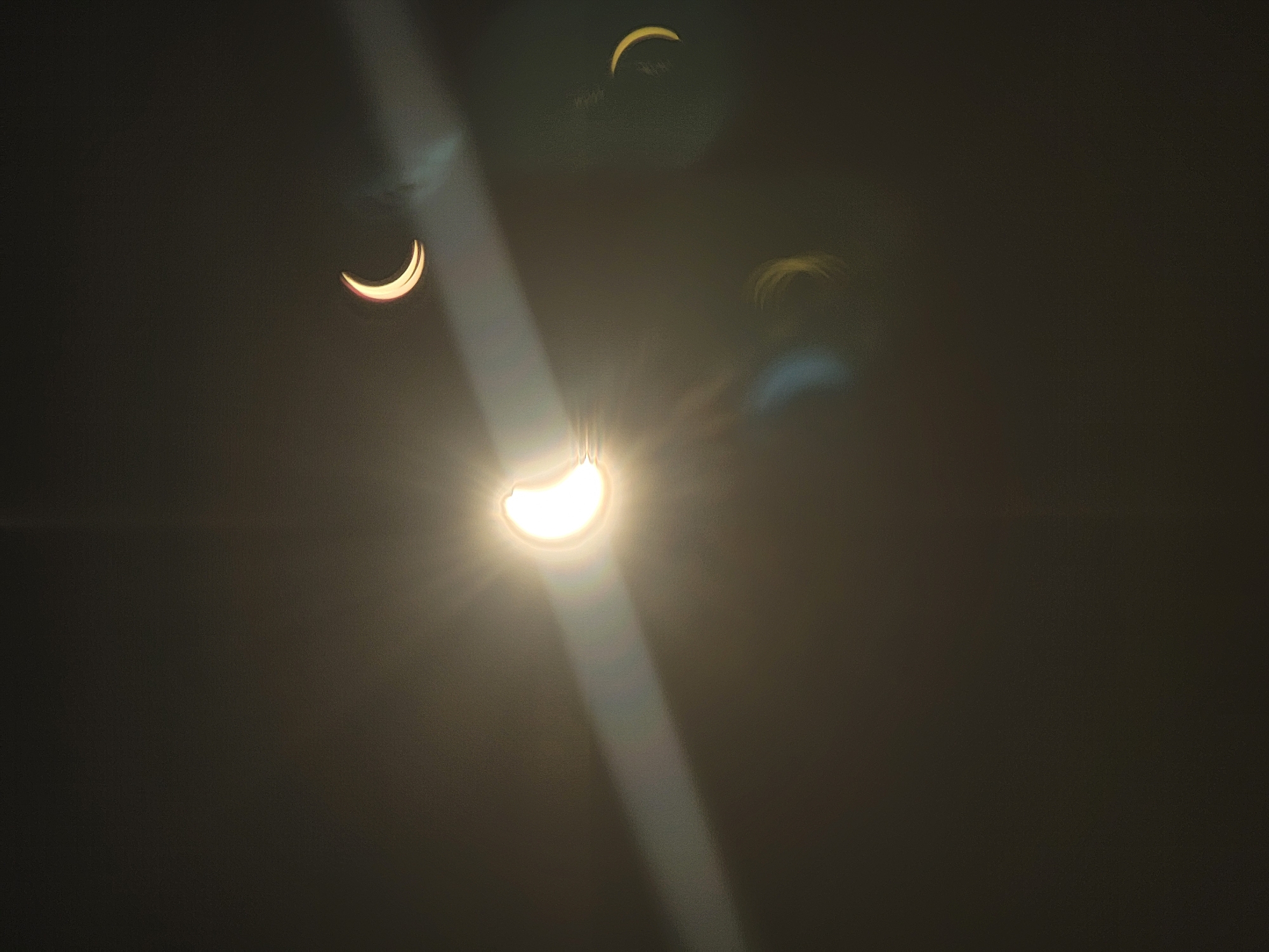 The image depicts a solar eclipse, with multiple crescent-shaped silhouettes of the sun visible against a dark, atmospheric background. A bright, elongated lens flare or light streak emanates from the left side of the frame, crossing diagonally towards the upper right. Three distinct crescent shapes are observable: one large and bright in the center left, another smaller and dimmer in the upper right, and a third, fainter crescent shape in the lower right. There are also some indistinct, possibly lens-flare related light artifacts in the lower portion of the image, appearing as soft, blurred streaks of yellow and blue. The overall ambiance is dark and ethereal, suggesting an astronomical event captured during twilight or at night. No people or specific urban features are identifiable, and thus, no connection to Quarteira, Portugal can be made from the visual information provided.