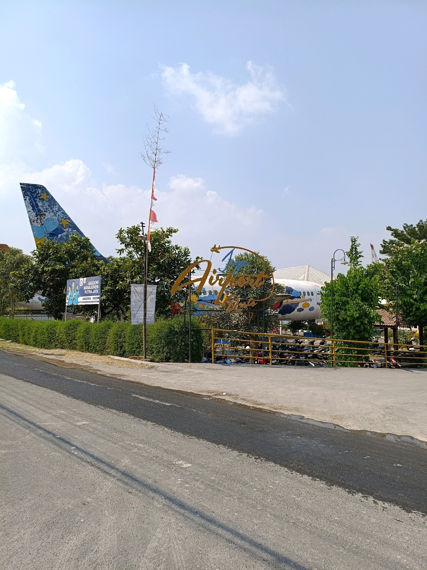 The image shows an abandoned airplane with the tail fin prominently displayed, painted with a blue and yellow design. The airplane is situated outdoors, surrounded by greenery and a paved area. A large, stylized sign with the words "Airport Views" is visible in the foreground, in front of the airplane. There are trees and bushes surrounding the airplane, and a yellow fence encloses part of the area. A paved road runs in front of the scene. A small group of motorcycles are parked behind the fence. The sky is blue with some white clouds. A flag with red and white stripes is flown from a pole near the entrance. There are also several signs and what appears to be a kiosk or shelter with a thatched roof in the background. The location is identified as Purwomartani, Indonesia, and the scene suggests a tourist attraction or a unique establishment built around the airplane.