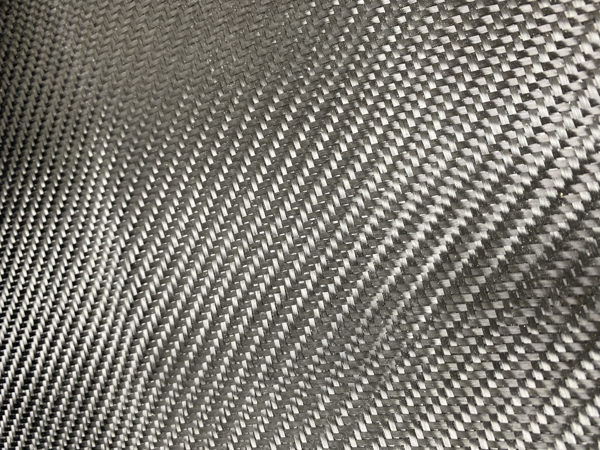 The image shows a close-up, high-angle view of a carbon fiber weave. The texture is a repeating diamond pattern of interwoven dark gray and black fibers. The fibers are thin and tightly packed, creating a textured, somewhat reflective surface. There are no people, objects, or identifiable landmarks present in the image to provide any context about its location in Vladimir, Russia.