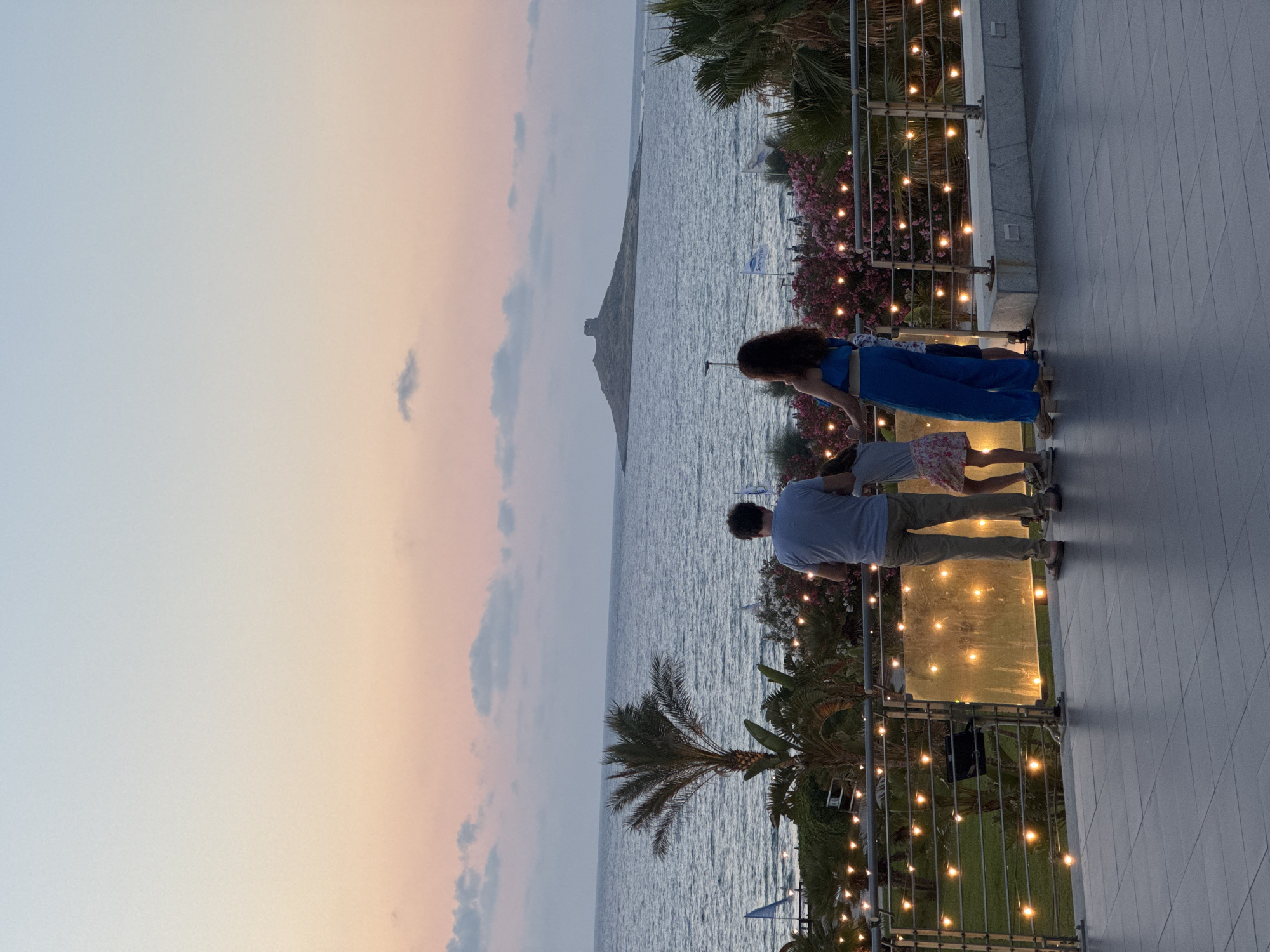 The image shows a family of three standing on a balcony overlooking the sea at dusk. The sky is a gradient of pale blue and pink, with some scattered clouds. A small island with a tower on it is visible in the distance. In the foreground, there are string lights illuminating the balcony, and palm trees and pink flowers are visible on the ground below. The family consists of a man, a woman, and a child. The man is wearing a blue t-shirt and khaki pants. The woman is wearing a blue jumpsuit. The child is wearing a pink and white dress. They are all looking out at the sea. The scene is peaceful and idyllic, capturing a moment of family togetherness. Isola delle Femmine is a small island town in Sicily, Italy, known for its beautiful coastline and charming atmosphere. The image appears to have been taken from a vantage point overlooking the town's marina or beach.