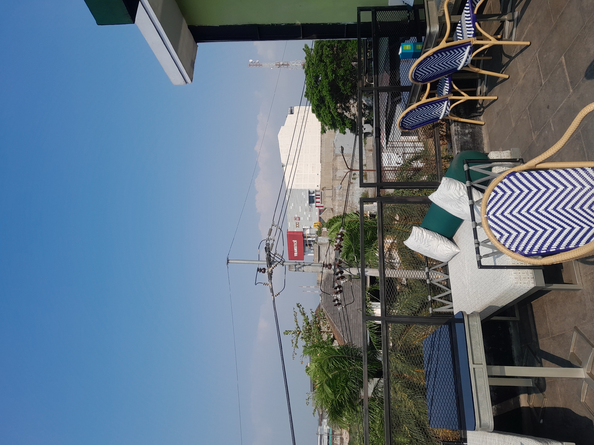 The image shows an outdoor seating area on a rooftop in Sempusari, Indonesia, under a clear blue sky. In the foreground, there is a modern outdoor lounge set with a white patterned couch and several cushions, as well as a table with a dark blue top. To the right, there are two rattan chairs with blue and white chevron patterned cushions. In the background, there are various buildings, including what appears to be a commercial building with a red sign that reads "TRANSMODERN", and utility poles with power lines stretching across the frame. Green foliage from trees is visible amidst the urban landscape. The overall scene depicts a relaxed, open-air environment in a developed urban area.