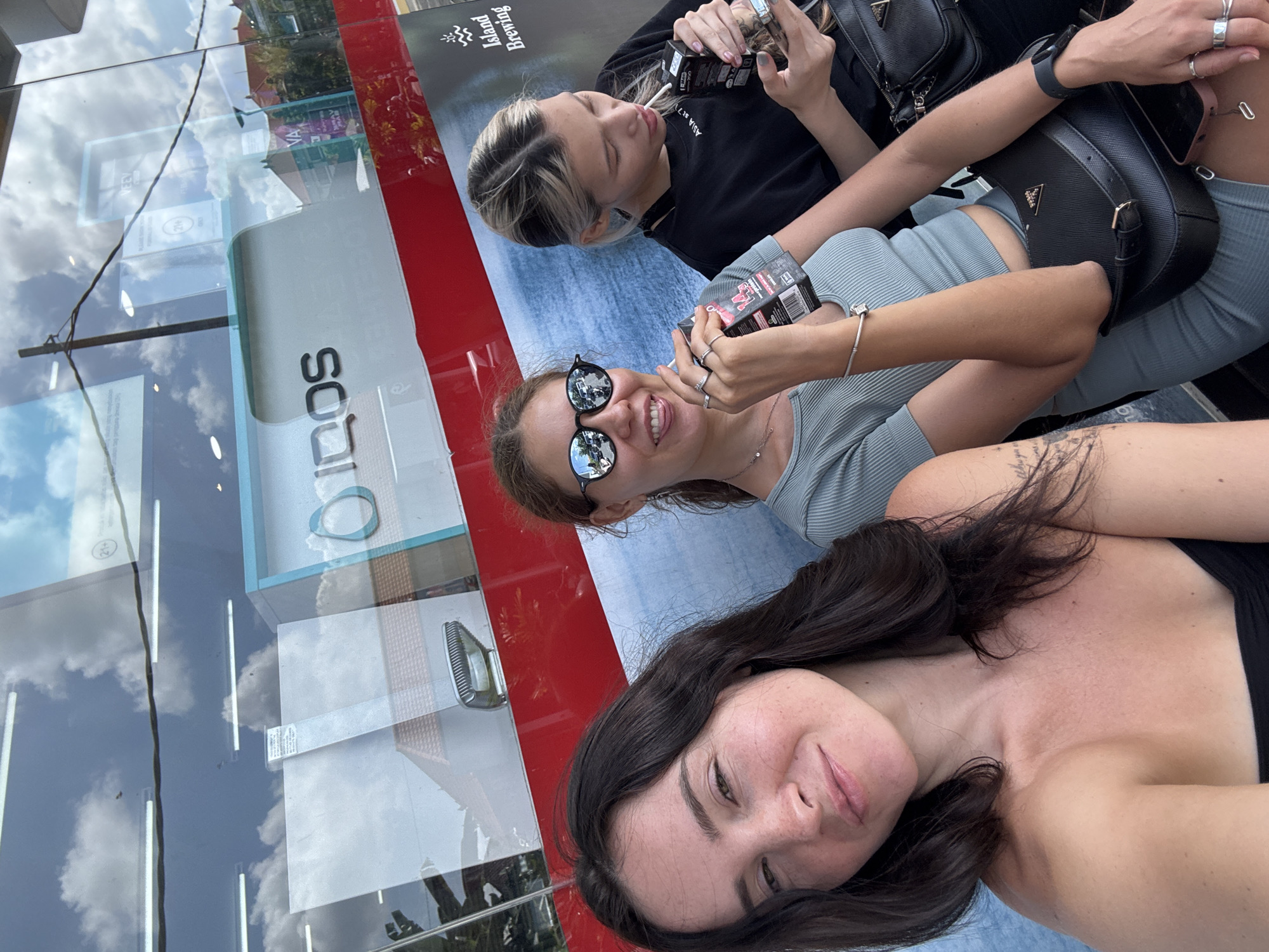 Three young women are posing for a selfie outdoors. The woman in the foreground, with dark hair, is smiling at the camera, her head tilted. She is wearing a black top. To her left, a woman with blonde hair is looking down, holding a small black box with a red label. She is wearing a grey ribbed top. Another woman, to the left of the blonde woman, is also looking down, holding a similar black box. She is wearing a grey top and black sunglasses. Both blonde women are wearing rings on their fingers and bracelets. Black handbags are visible, one on the blonde woman's shoulder and another on the grey-topped woman's lap.
The background appears to be a street scene with reflections on glass. A red and white striped surface is prominent. In the reflections, clouds are visible, suggesting it is daytime with clear skies. Text visible on a poster in the background is "Island Brewing". The logo "iqos" is also visible on a panel. The scene is set in North Kuta, Indonesia. The women appear relaxed and are engaged in a casual activity, possibly shopping or taking a break.