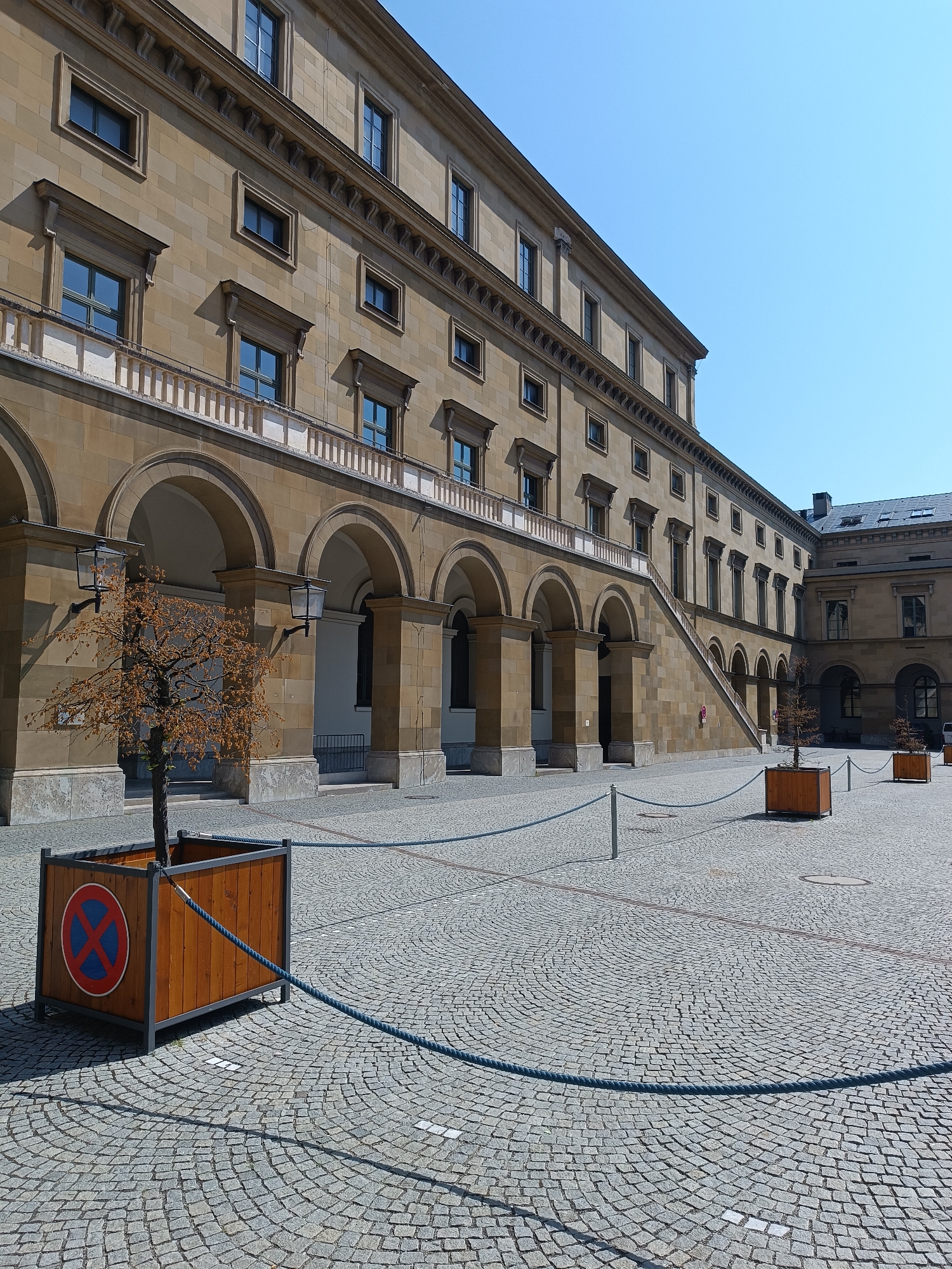 The image depicts the courtyard of the Residenz in Munich, Germany. The courtyard is paved with cobblestones arranged in a circular pattern. To the left, a long, multi-story sandstone building with arched colonnades and a balcony runs along the courtyard. A bare-branched tree in a wooden planter with a "no parking" sign is located in the foreground. Blue ropes are strung between metal posts, cordoning off areas of the courtyard. In the background, another building with a pitched roof and multiple windows is visible. The sky is clear and blue.
