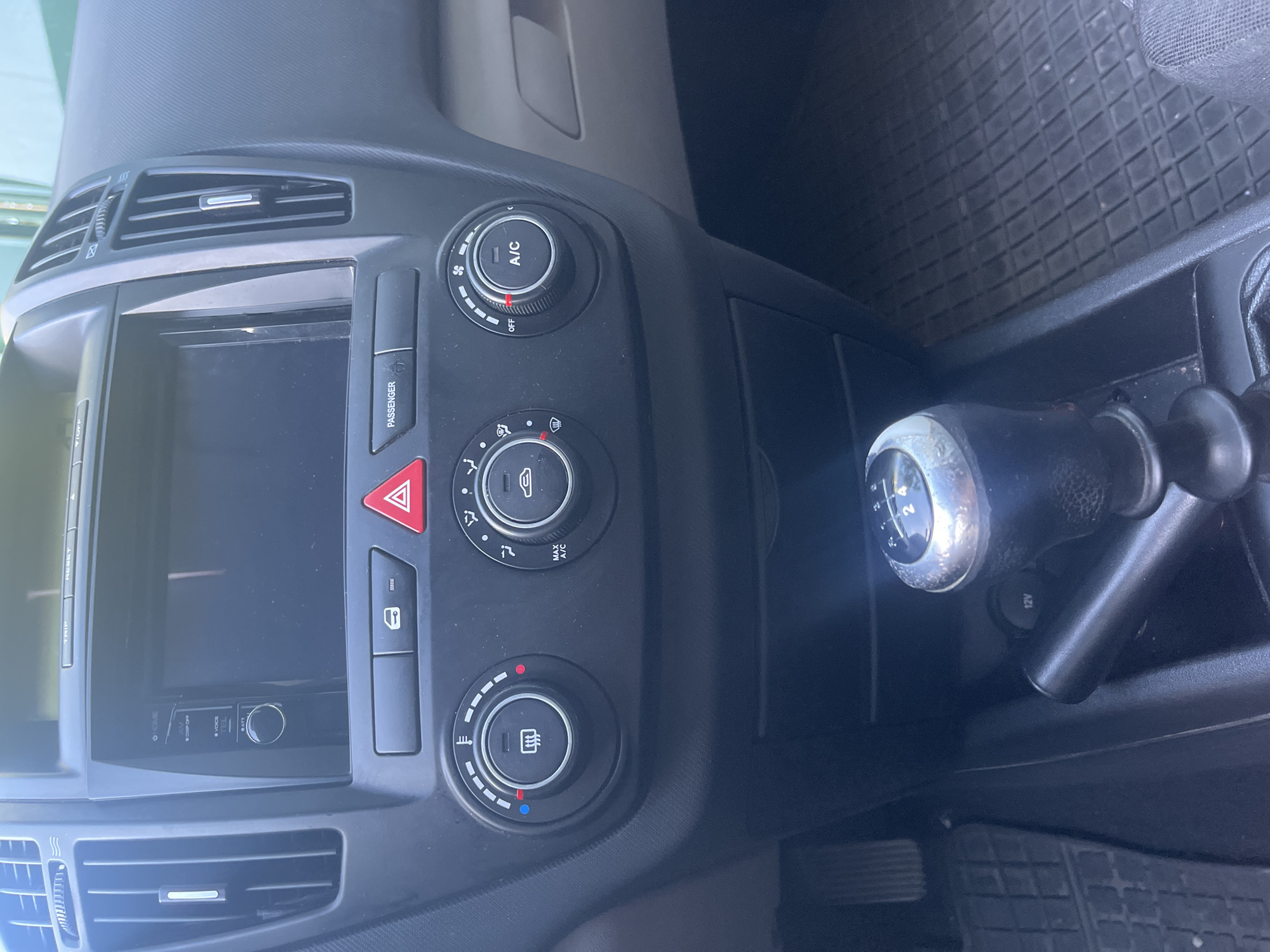 The image shows the interior of a car, specifically the center console area. Visible features include the car's infotainment screen, climate control knobs, a hazard light button, a passenger airbag indicator, and a gear stick, indicating it is a manual transmission vehicle. A 12V power outlet is also visible near the gear stick. The floor mats are black with a grid pattern. The environment suggests the interior of a car, and there's no information in the image related to Blagoevgrad, Bulgaria.