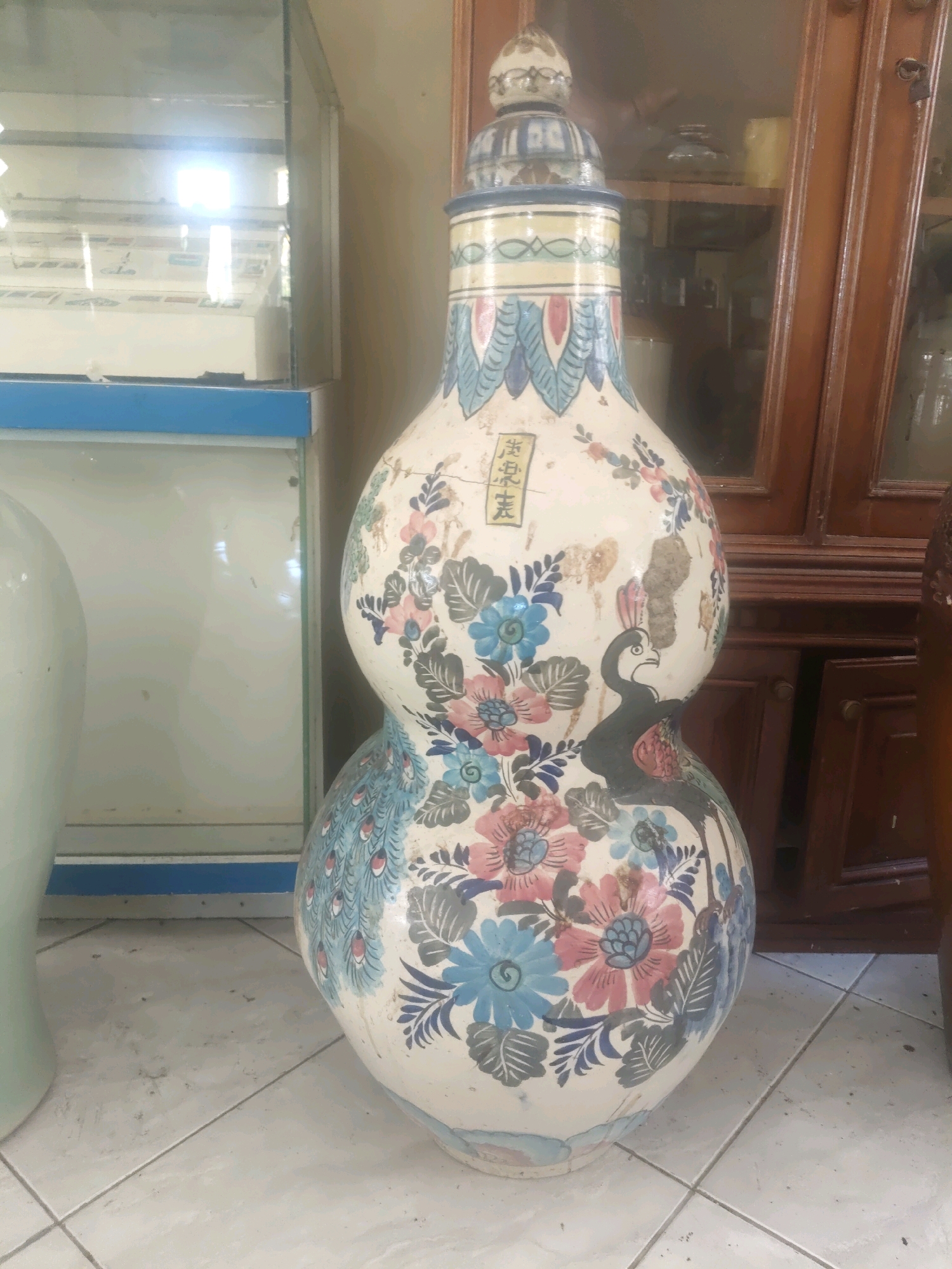 The image shows a large, ornate, gourd-shaped ceramic vase decorated with floral patterns and peacocks. The vase has a white base with blue, pink, and green floral motifs, and several depictions of black and white peacocks. The vase is topped with a decorative lid. To the left of the vase is a portion of a light-colored, smooth-surfaced object, possibly another vase or pedestal. Behind the vase and to the right, there is a dark wooden cabinet with glass doors, revealing shelves and items inside. The floor is tiled with off-white square tiles. To the far left, a glass-fronted display case with blue trim is visible. The lighting suggests an indoor environment, potentially a shop or storage area. There are no people visible in the image. The location is identified as Cisaga, Indonesia.