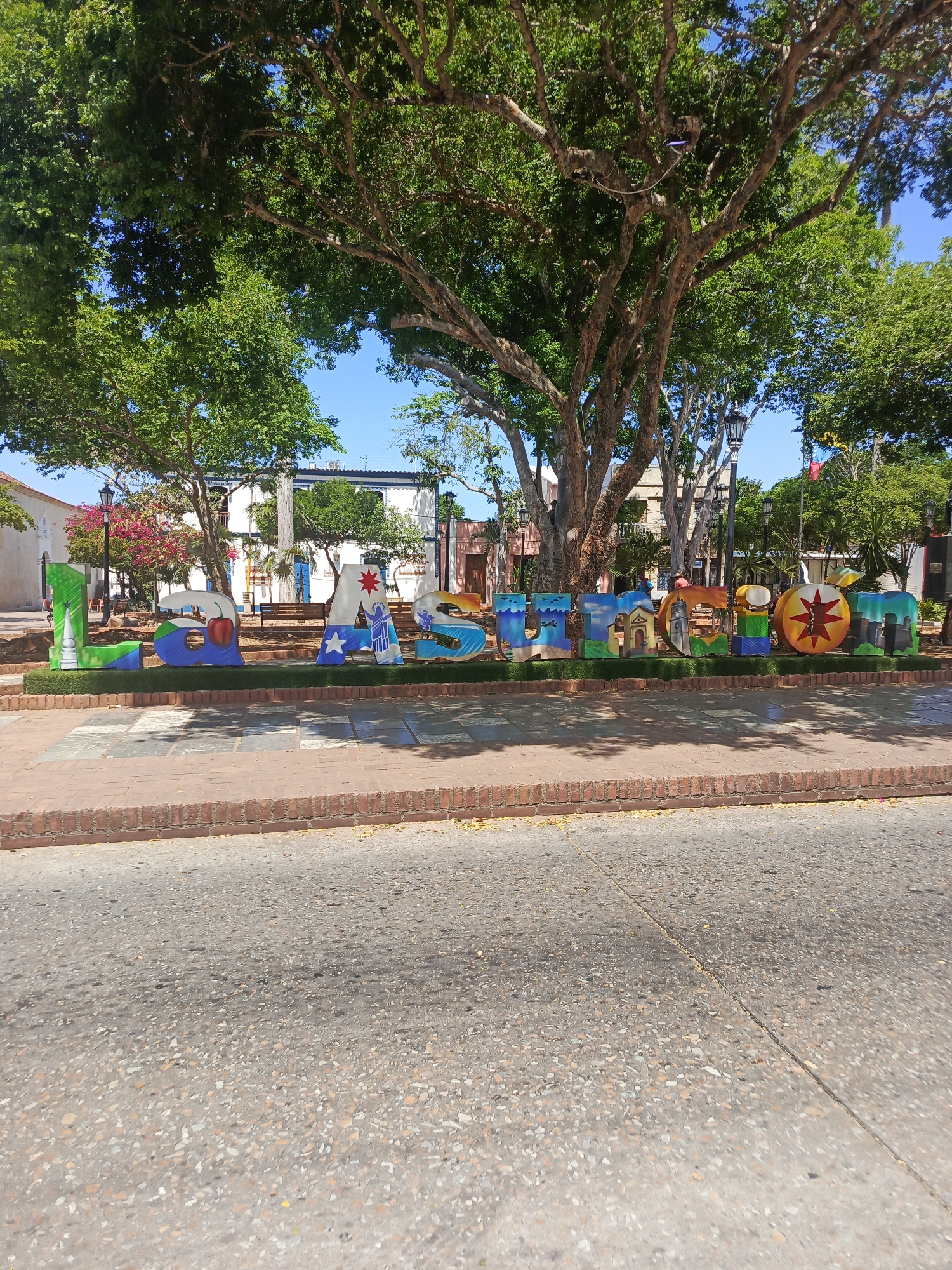The image shows a street view in La Asuncion, Venezuela. A sign spelling out "LA ASUNCION" is displayed prominently in the foreground. The sign is colorful and appears to be made of individual letter-shaped structures, each decorated with different images. Behind the sign, there is a public square or park with trees and buildings. The scene is well-lit, suggesting it is daytime. The street in the foreground is paved and appears to be in good condition. There are no people immediately visible in the image.