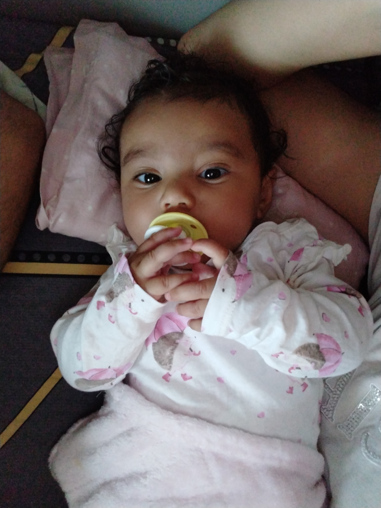 A baby with curly dark hair and brown eyes lies on their back on a pink pillow. They are wearing a white long-sleeved shirt with pink umbrellas and hedgehogs on it, and they have a yellow pacifier in their mouth, which they are holding with their hands. The baby is also partially covered by a pink blanket. The background shows a dark grey and brown surface. The location is unknown.