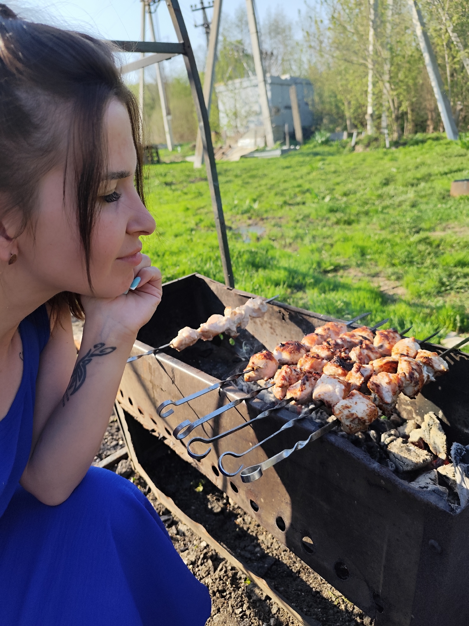 A woman is sitting next to a barbeque grill, with skewers of meat cooking over hot coals. The woman is looking off to the side, with her chin resting on her hand. She has dark hair pulled back and is wearing a blue dress. A tattoo is visible on her wrist. The barbeque grill is made of dark metal and has several skewers of meat on it. The meat appears to be chicken and is browning. In the background, there is a grassy area with some trees and a building. There are power lines on poles in the background. The location is likely in the city of Фоки, Russia.