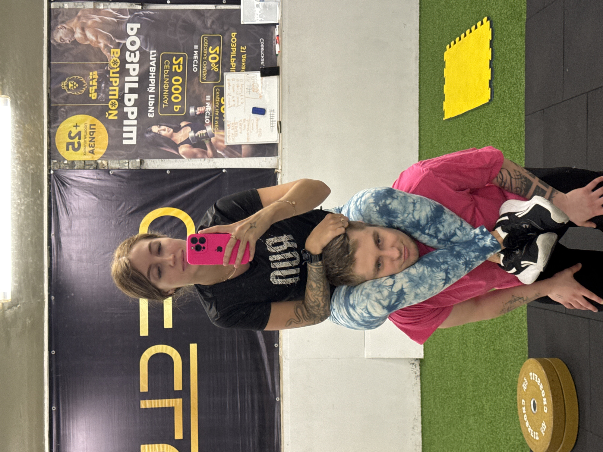 **Scene Description:**
The image shows a selfie taken inside a fitness facility in Chaikovsky, Russia. A woman is holding a bright pink mobile phone and taking the selfie with a man. The man is being put in a submission hold. They are both wearing workout clothes.
**Environment:**
Behind them, there is a fitness facility with advertisements for fitness services written in Cyrillic. A wall in the background has gym puzzle mats. A weight plate is on the floor. A white board has markings on it.
**People:**
* A woman holding a pink mobile phone.
* A man being put in a headlock.
**Actions/Interactions:**
* A woman taking a selfie.
* A man being put in a submission hold.
**Objects:**
* Mobile phone
* Weight Plate
* Gym advertising
* Puzzle mats
* White board
* Workout clothing