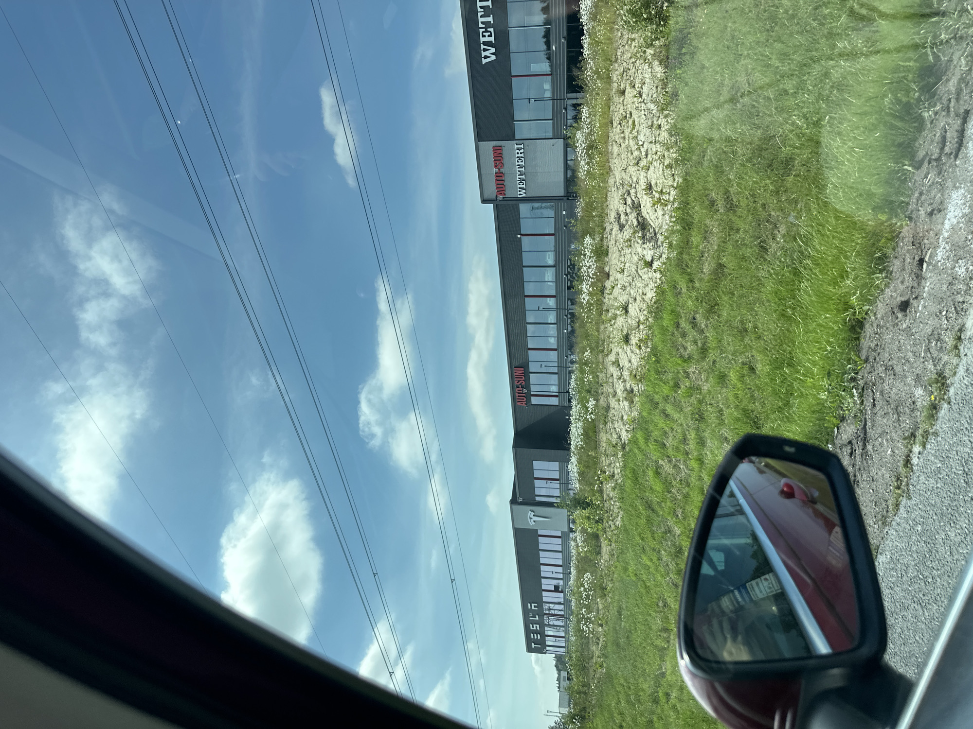 A long, dark-colored commercial building is visible under a bright blue sky with scattered white clouds. The building has a modern facade with many large windows. Across the front of the building, there are several signs. The leftmost sign reads "TESLA" in white, capitalized letters, with a red Tesla logo to its right. To the right of that, a sign reads "AUTO-SUNI" in red, capitalized letters, and below that, "WETTERI" in smaller red letters. Further to the right, another sign reads "WETTERI" in white, capitalized letters. A grassy area with some scattered white wildflowers is in front of the building. In the foreground, a portion of a red car is visible, including its side mirror, which reflects the grassy area and the building. The car appears to be in motion, as suggested by the perspective and the reflection. Power lines are strung across the sky above the building.
