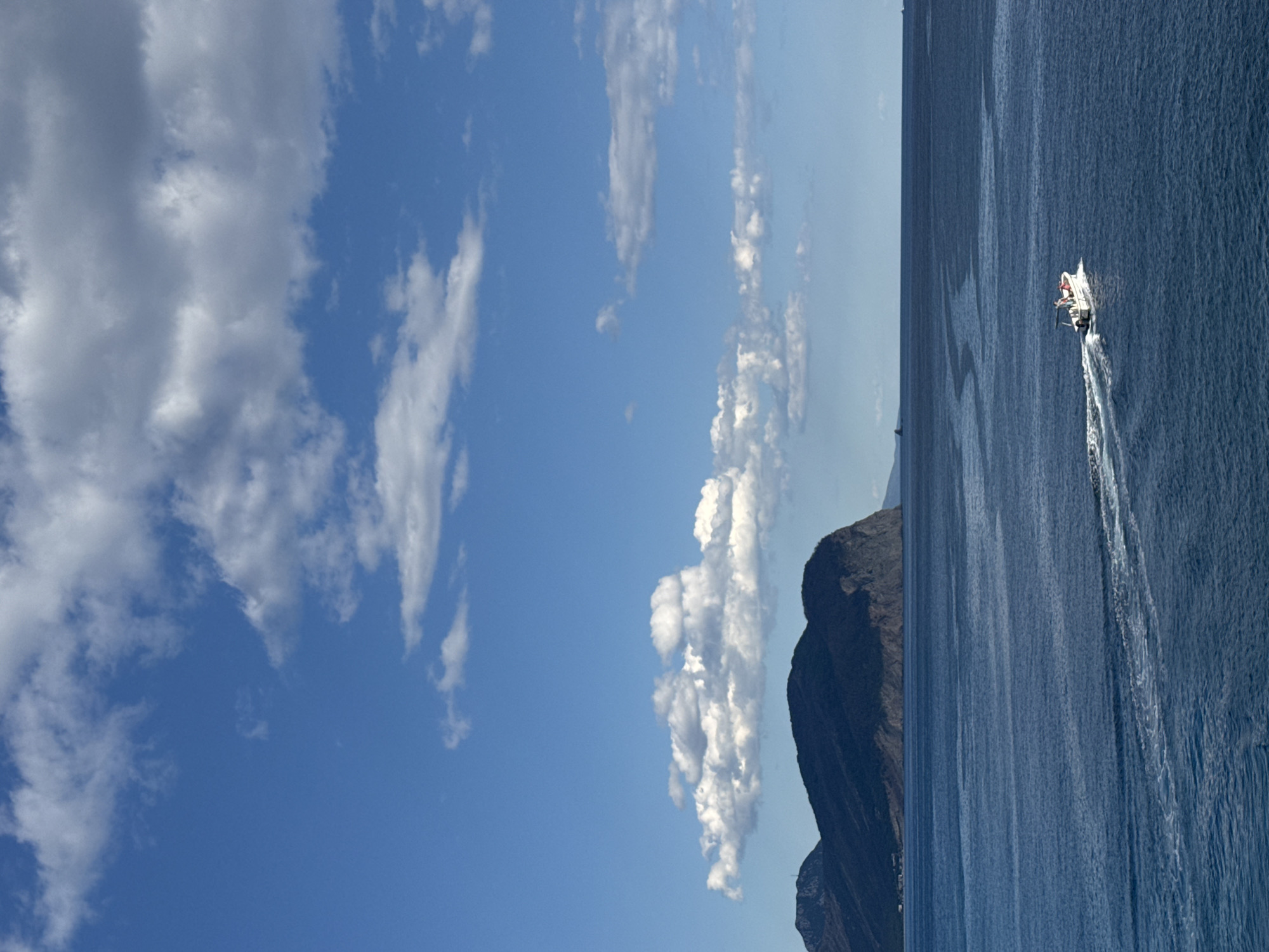 A wide-angle, high-angle shot captures a vibrant blue sky dotted with white, fluffy clouds. Below, a vast expanse of deep blue water stretches towards a rugged, dark mountain range that dominates the horizon on the left. In the foreground, a small white boat with a red trim is traversing the water, leaving a distinct white wake behind it. The boat appears to have at least one person on board. The scene is likely Petrovac centar, Montenegro, given the characteristic mountainous coastline.