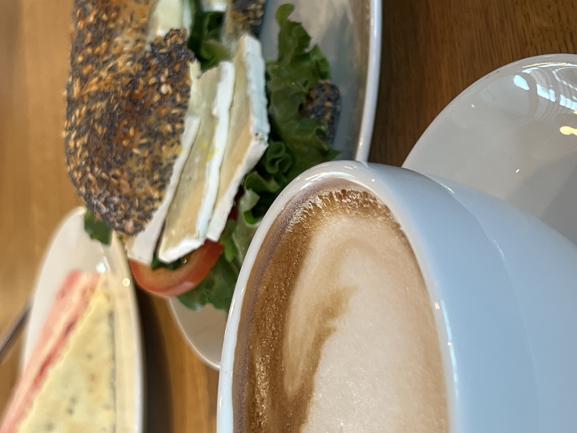 A close-up shot shows a cup of latte with frothy milk and a dark coffee swirl in the foreground, placed on a saucer. In the background, a plate holds a halved bagel sandwich with a variety of toppings, including lettuce, tomato, and slices of white cheese, topped with a sprinkle of seeds. Another plate, blurred and out of focus, is visible to the left, with a slice of cake featuring pink layers. The scene is set on a wooden table, suggesting a cafe or restaurant environment, possibly in Moosburg, Germany.