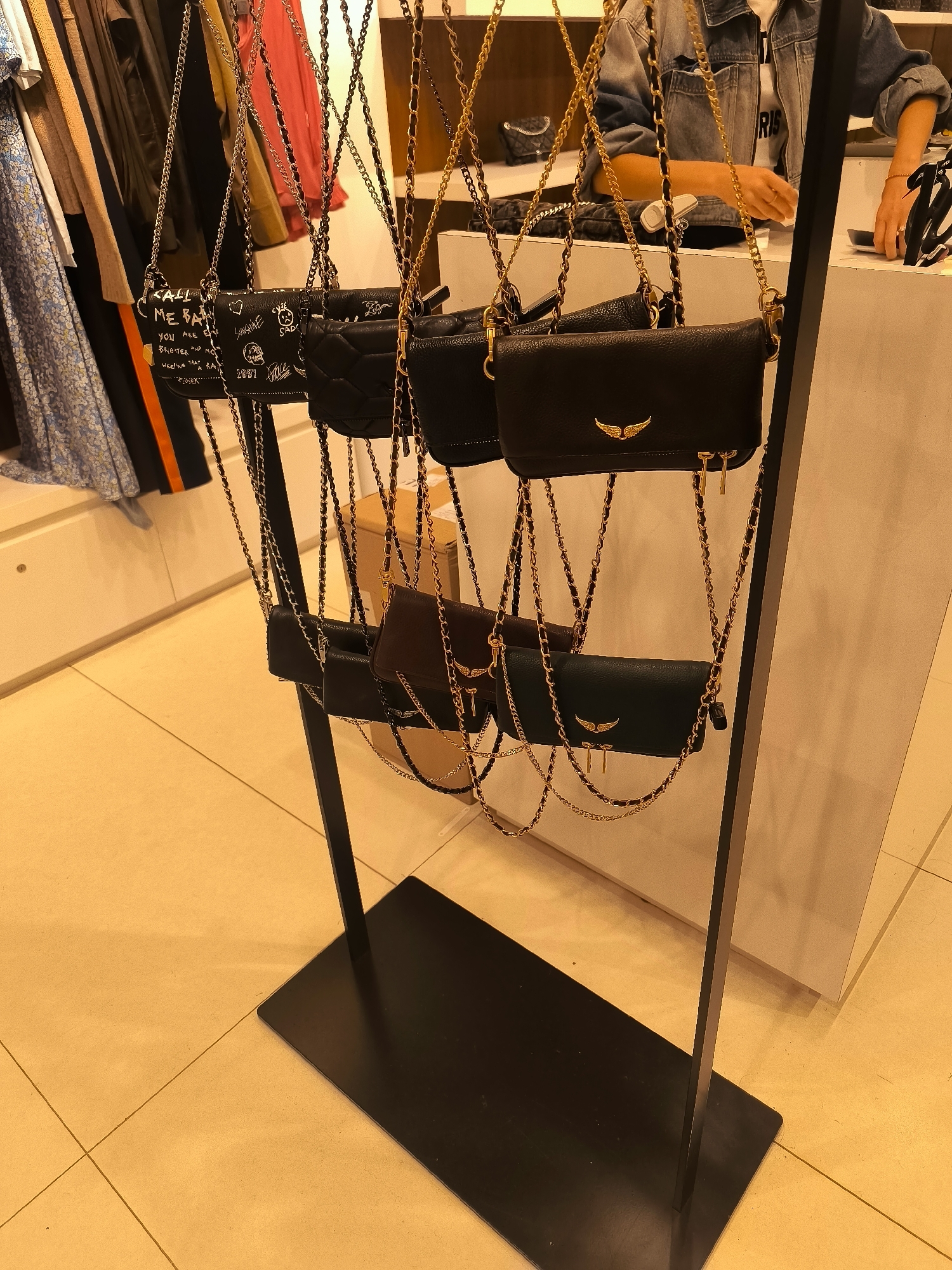 The image shows a retail display of handbags in a store. Several black leather handbags with gold chain straps are hanging from a black metal rack. Some of the handbags have decorative stitching, while others have written designs. In the background, clothing items are visible on hangers, and a person wearing a denim jacket is interacting with a sales counter. The floor is tiled in a light yellow color, and the lighting suggests an indoor environment. The scene is located in a retail store, likely a fashion boutique, in The Hague, Netherlands.
