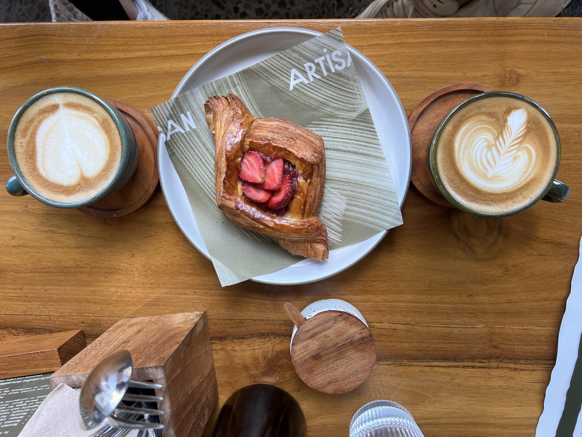 The image displays a flat lay of a wooden table with several items, suggesting a cafe setting in Mengwi, Indonesia. Two cups of latte art coffee are present, one with a heart shape and the other with a leaf pattern. A pastry with fresh strawberries sits on a white plate, accompanied by a paper napkin with the text "ARTISAN" visible. To the left, a wooden cutlery holder contains forks and spoons, and a small ceramic container with a wooden lid is also visible. The overall ambiance is relaxed and indicative of a pleasant cafe experience.