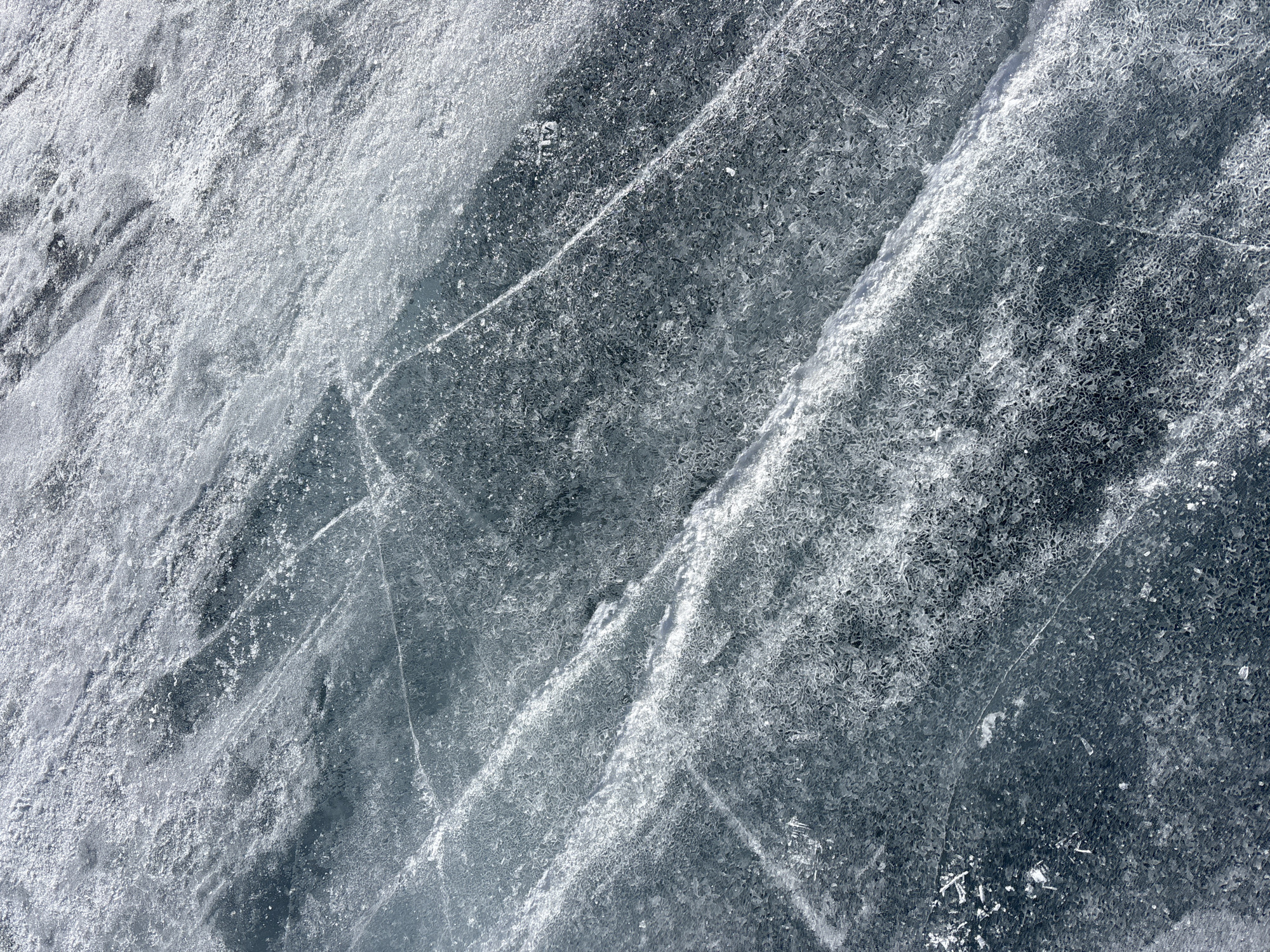 The image displays a close-up, aerial perspective of a textured ice surface located in Listvyanka, Russia, presumably on Lake Baikal. The scene is devoid of people, subjects, or explicit actions beyond environmental interactions with the ice. The primary object is a large expanse of frozen water, characterized by a grey-blue base tone. Numerous linear fractures and cracks, varying in width and orientation, crisscross the surface, appearing as lighter, opaque lines. Irregular patches of frosted or granulated ice crystals are distributed across the scene, creating areas of heightened reflectivity and a coarse texture. Segments of clearer, smoother ice are interspersed with these frosted regions. In the upper right quadrant, a faint, three-letter inscription, appearing as "TLV" or "TLY," is etched into the ice surface, indicating human interaction. The overall environment is illuminated, suggesting natural daylight on an extensive outdoor ice formation.