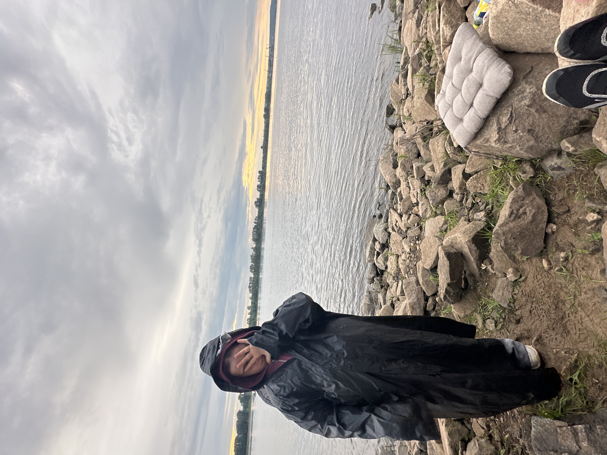 A person wearing a black hooded raincoat and a maroon sweatshirt covers their face with their hand. They are standing on a rocky shoreline next to a wide body of water. The sky is overcast with some patches of yellow light from the setting sun visible on the horizon. In the foreground, there are several large rocks, some with green grass growing between them. A light-colored cushion sits on a rock to the right of the person, and a pair of black and white sneakers are visible in the bottom right corner of the frame. The location appears to be a natural setting, likely a riverbank or lake shore.
