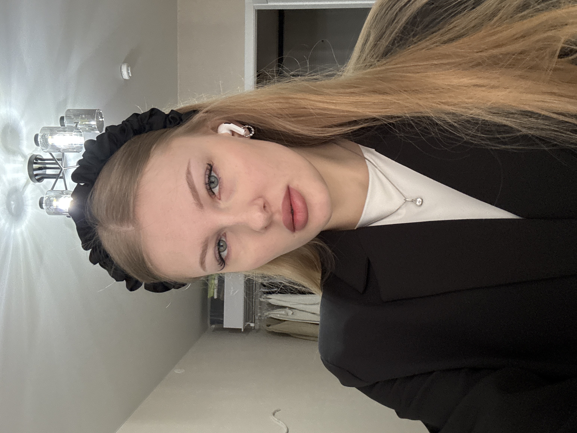 A young woman with fair skin, blonde hair, and blue eyes is photographed indoors, likely in Perm, Russia given the request for location specificity. She is wearing a black blazer over a white shirt with a silver necklace and has a black scrunchie headband. She also has wireless earphones in her left ear. The background includes a modern-style chandelier, a door, and what appears to be a desk area with items such as paper and shelves. The lighting is artificial. The woman is looking at the camera.