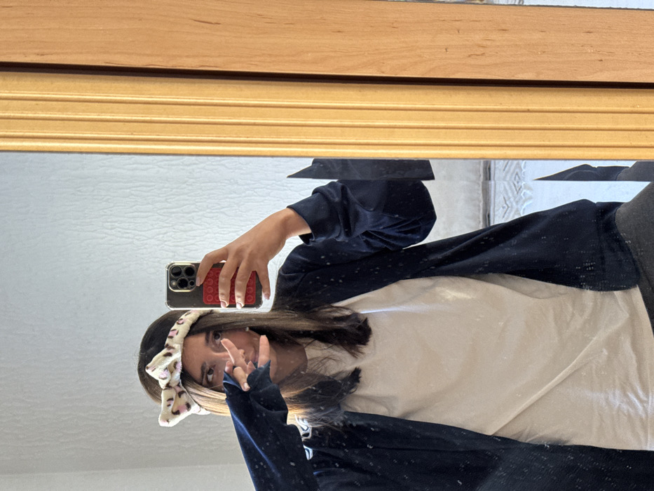 An individual is depicted taking a mirror selfie indoors in Zankovići, Montenegro. The subject, a female, has long, straight dark hair and wears a cream-colored headband with a pink and brown leopard print, tied in a bow. She is dressed in a white t-shirt and a dark blue open cardigan. Her right hand holds a silver smartphone with a clear case and a red grid-patterned pop-socket, oriented vertically to capture the reflection. Her left hand is raised, with fingers forming a 'peace' sign or similar gesture, partially obscuring the lower half of her face. The reflection shows a textured white wall in the background and a partial view of a light-colored surface or object at the bottom. The mirror is framed on the right side by a golden, fluted decorative trim adjacent to a plain light-wood panel.