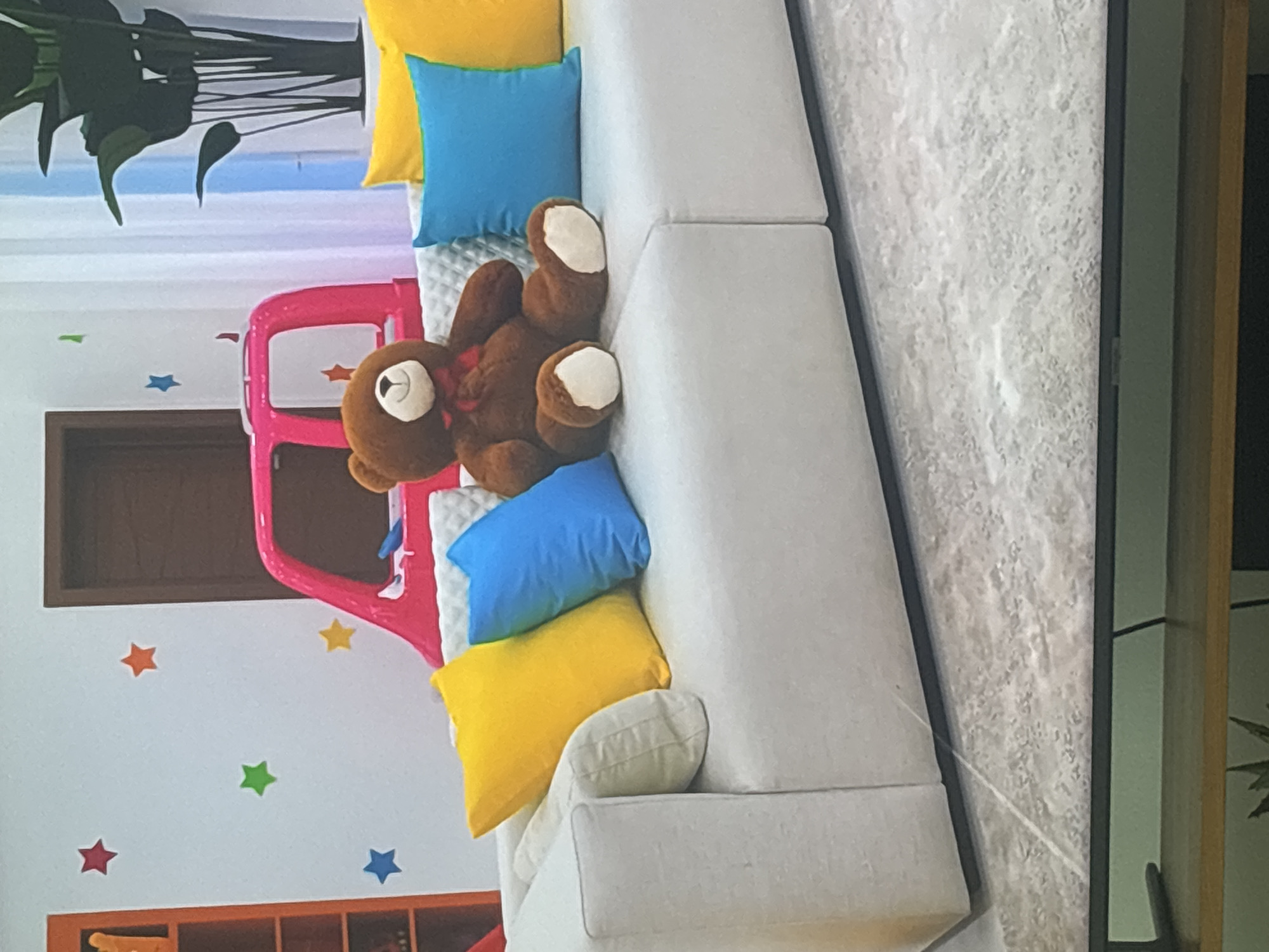 A brown teddy bear with a red ribbon around its neck sits on a white sectional sofa. The sofa is adorned with several colorful pillows, including yellow and blue. In the background, a pink toy car is visible, along with a tall green plant and a white curtain. The walls are decorated with multi-colored star decals. The scene appears to be indoors, possibly in a child's playroom or a modern living space. There is no indication that this image is located in Dubai, United Arab Emirates.