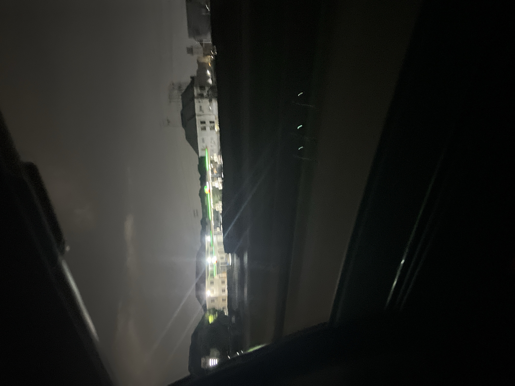 The image is taken from inside a vehicle at night, possibly Gwarinpa, Nigeria, facing outwards. The scene includes the street lights on, tall buildings, and a hazy sky.