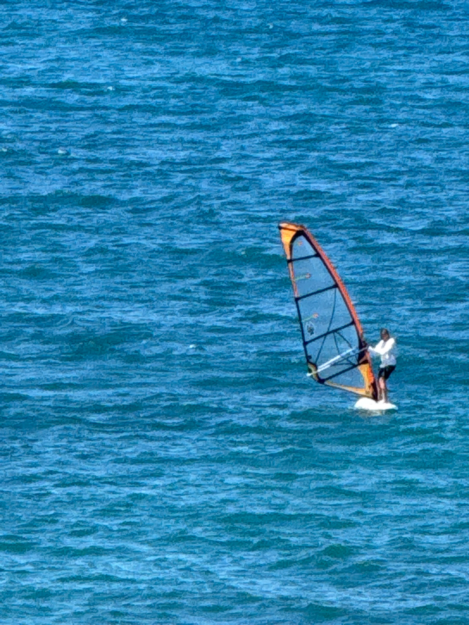 A person is windsurfing on the bright blue, choppy water. The windsurfer is wearing a white long-sleeved shirt and dark shorts, standing on a white board. The sail of the windsurf board is triangular and orange and blue with black struts. The windsurfer is positioned to the right of the image. The image captures a sunny day with a vast expanse of water. The location is the Black Sea near Anapa, Russia.