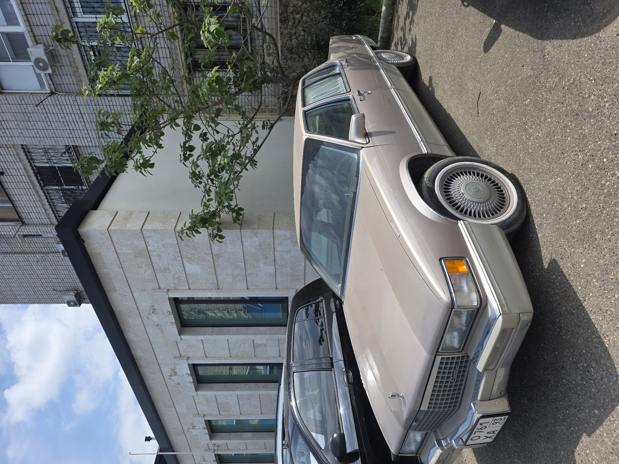 The image shows a tan Cadillac parked on a street in Krasnodar, Russia. The car is parked next to a modern building with a light-colored facade. A black car is parked behind the Cadillac. The license plate of the Cadillac is visible, reading "O 761 XA 93". A tree is growing near the building, partially obscuring it. The sky is visible in the upper left corner of the image.