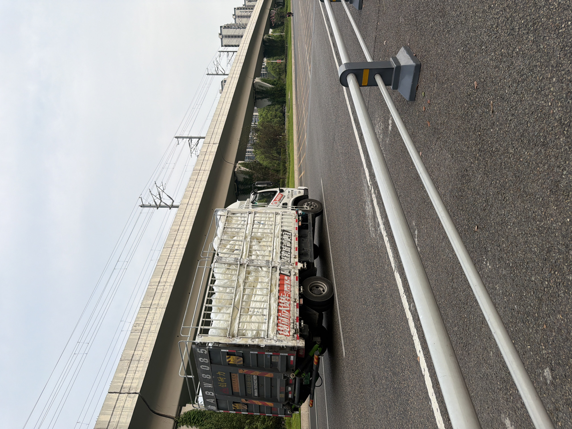 A truck with a white cargo load is driving on a paved road in Jiangjin, China. The truck is dark gray with a white cab and has a license plate reading "GA 8939Z". The cargo is covered in white plastic. In the background, an elevated concrete structure, likely a bridge or overpass for a railway, is visible. Overhead power lines run along the structure. Trees and modern buildings are also present in the background, suggesting an urban or suburban environment. A motorcycle is seen in the distance on the right side of the road. A white metal railing with dark gray posts lines the right side of the road. The sky is overcast.