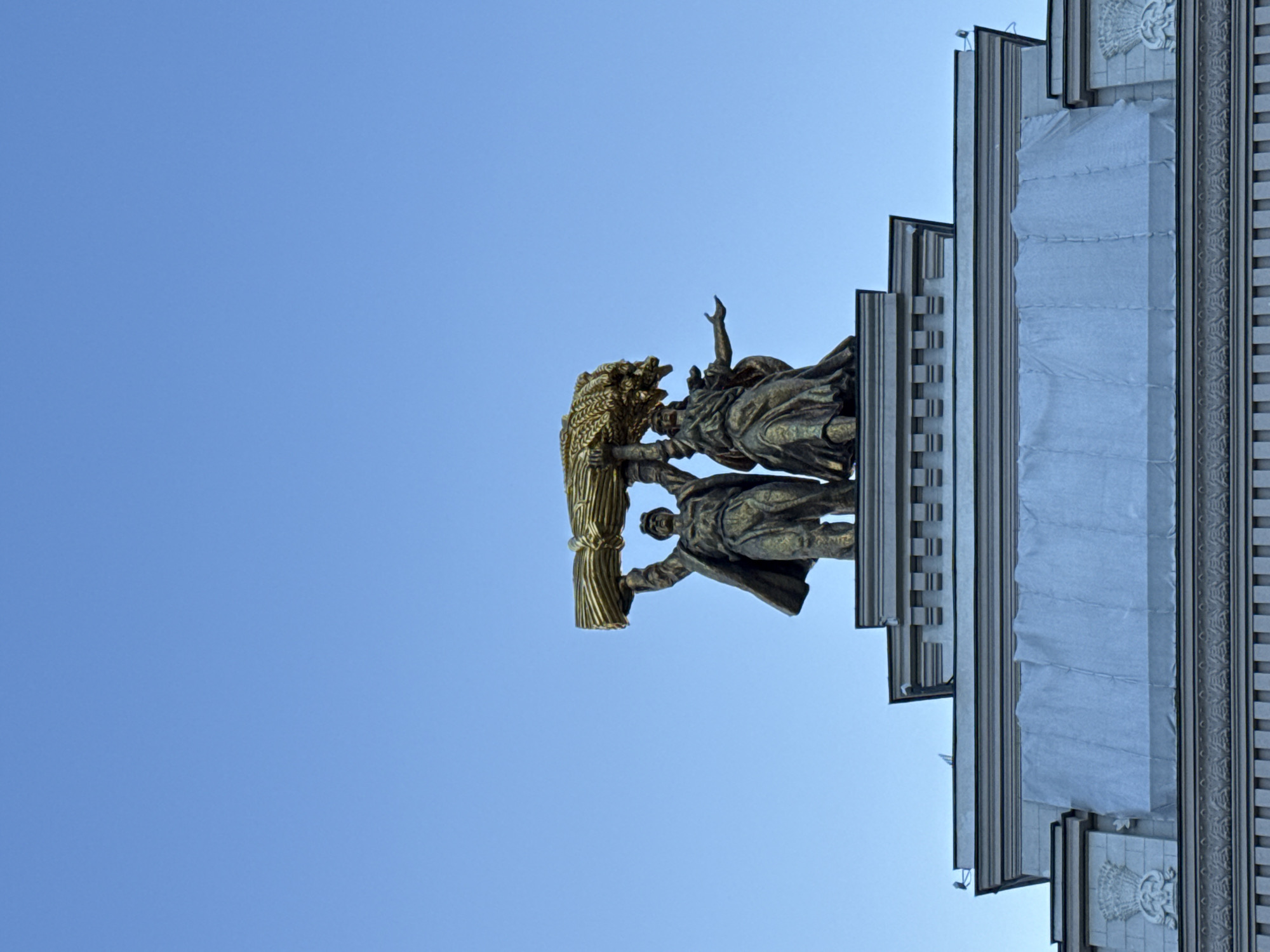 The image captures a low-angle view of the "Worker and Kolkhoz Woman" monument, a gilded bronze sculpture, against a clear blue sky. The monument depicts a male and female figure holding aloft a sheaf of wheat. They stand on a tiered structure, part of a larger building. The building's facade is adorned with architectural details, including relief sculptures and what appears to be a light-colored tarp covering a section of its upper portion. The monument is situated on Prospect Mira in Moscow, Russia, and is a prominent landmark of the city.