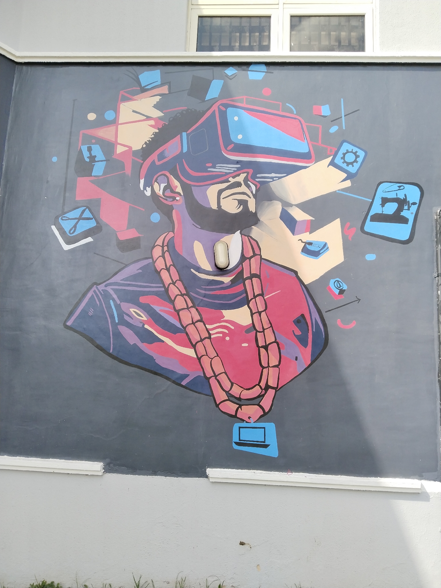 A mural is painted on an exterior wall, likely located in Uyo, Nigeria. The mural features a person wearing a virtual reality headset, with a thick chain necklace. Stylized images representing various activities or applications are scattered around the person's head. These include scissors, a sewing machine, a gear, and a laptop. The wall is painted gray around the mural, with white trim along the top and bottom. Above the mural is a window. Grass and dirt are visible at the bottom of the image.