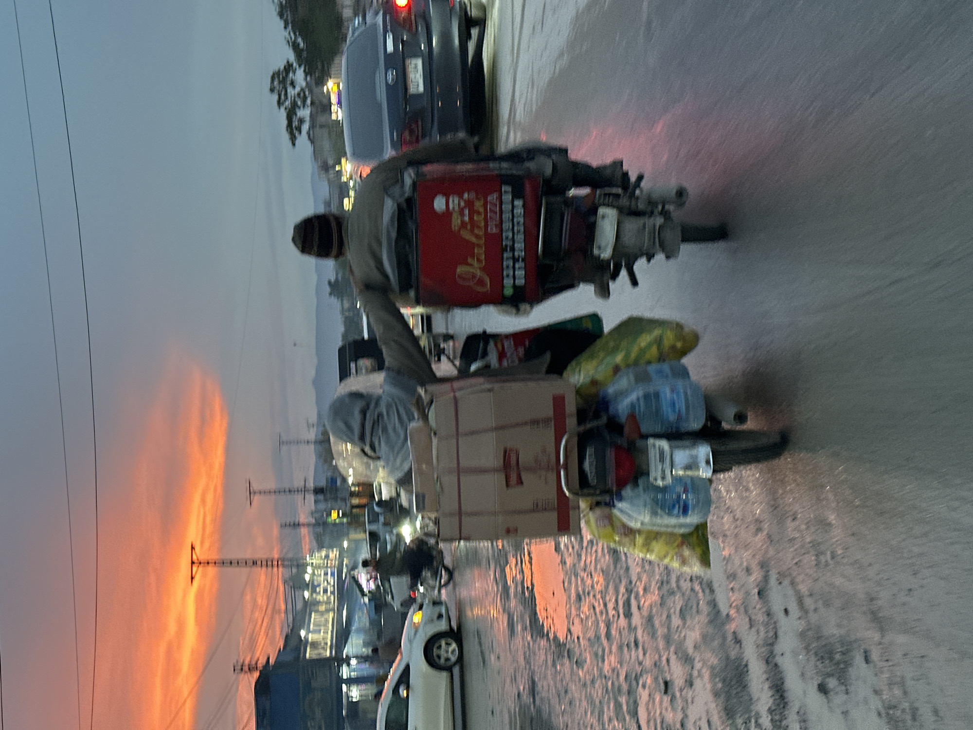 The image shows a man on a motorcycle in Quetta City Tehsil, Pakistan. He is delivering pizza, as evidenced by the "Italian Pizza" delivery box attached to the back of the motorcycle. The motorcycle is heavily loaded with goods, including boxes, water bottles, and other items. The background includes a city street with power lines, buildings, and a sunset in the distance. The road surface appears wet, reflecting the colors of the sky. Other vehicles are also visible in the scene, including a car in front of the motorcycle and another behind it.
