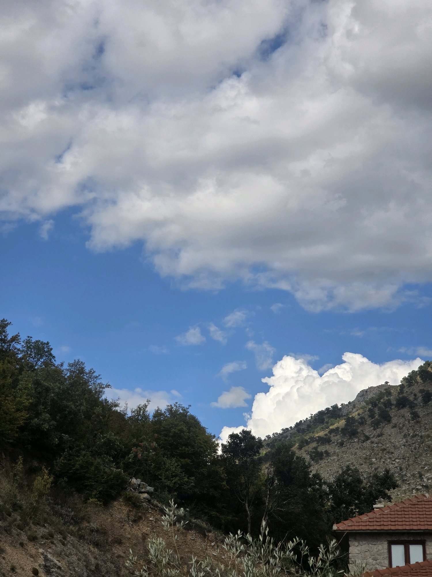 The image depicts a mountainous landscape under a partly cloudy sky. The foreground features a steep, rocky slope covered in dense green foliage and scattered dry vegetation. To the right, a portion of a stone building with a red tiled roof is visible. The middle ground shows more trees and a rocky hillside ascending towards the background. The background is dominated by a vast expanse of sky, with large, white, fluffy clouds interspersed with patches of bright blue. The scene appears to be located in a rural or semi-rural mountainous area, likely on the outskirts of Podgorica, Montenegro, given the characteristic Mediterranean vegetation and building style. There are no people or animals visible in the image.