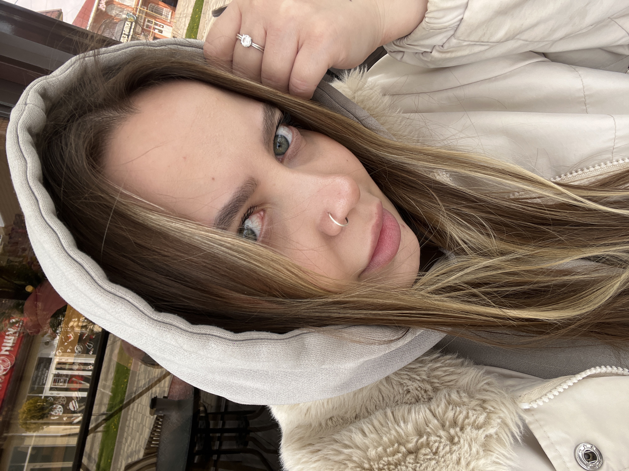 The image is a selfie of a woman with light brown hair and green eyes, wearing a light-colored hooded jacket with a fur lining. She has a nose ring and a ring on her finger. The photo is taken at an angle, and the background shows a reflection of a building in Novopokrovskaya, Russia. The building has a red sign with Cyrillic writing on it and appears to be a store. The reflection also includes trees, a sidewalk, and other buildings.