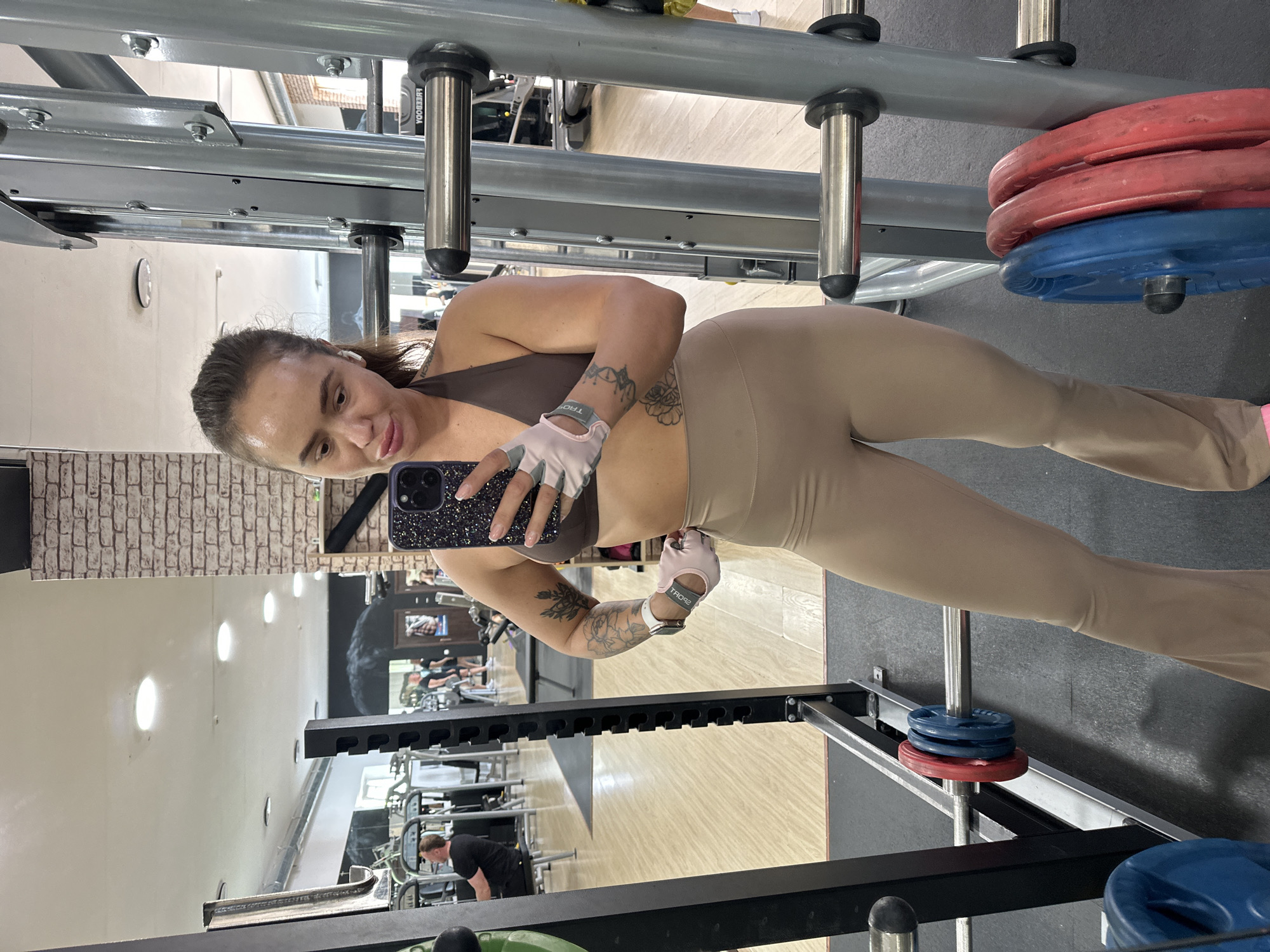A woman with long dark hair tied back and makeup poses for a selfie in a gym. She is wearing a brown halter-neck sports bra, beige flared leggings, and light pink workout gloves. She has tattoos on her arms. The gym features exposed brick walls, mirrors, weightlifting equipment, and weight plates. In the background, other gym-goers are visible. The gym appears to be located in Yuzhnouralsk, Russia.