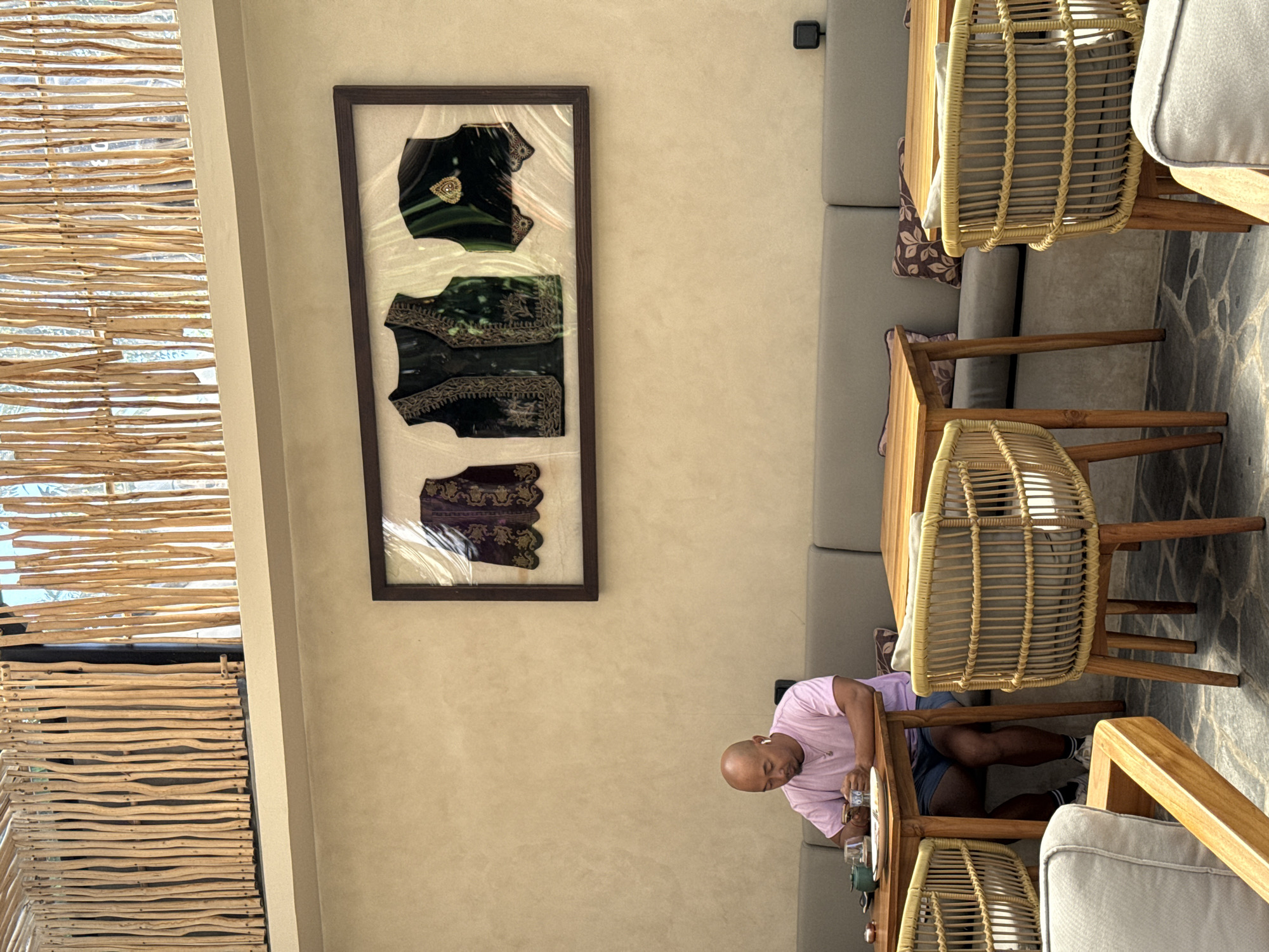 The image captures an indoor dining or lounge area, likely in a cafe or restaurant, presented in a portrait orientation. The scene is well-lit, suggesting daytime.
In the foreground and central-right, a man with a shaved head is seated at a wooden table. He is wearing a light pink t-shirt and dark shorts, and has white earbuds in his ears. He is looking down at an object on the table, possibly a mobile phone, and appears to be holding a clear glass with a drink. His legs are visible under the table.
The furniture includes several light wooden tables and chairs made of woven rattan with light grey cushions, characteristic of a natural, tropical aesthetic. Along the right wall, there's a long banquette sofa with light grey upholstery and patterned throw pillows. The floor is covered with large, irregularly shaped dark stone tiles.
On the left side of the frame, a textured wall screen made of horizontal bamboo or natural wood strips allows dappled light to filter through, hinting at an outdoor connection or window. On the main wall, above the man, hangs a large, rectangular framed artwork. This piece features three intricately designed, dark-colored garments or textile pieces, possibly traditional vests, displayed against a light background. Each piece has distinct embroidery and detailing.
The walls are a neutral, light beige color. The overall atmosphere appears relaxed and casually elegant, with a focus on natural materials. No visible text is clearly legible in the image. Given the context, this setting is located in the Ubud District, Indonesia, aligning with the tropical design elements.