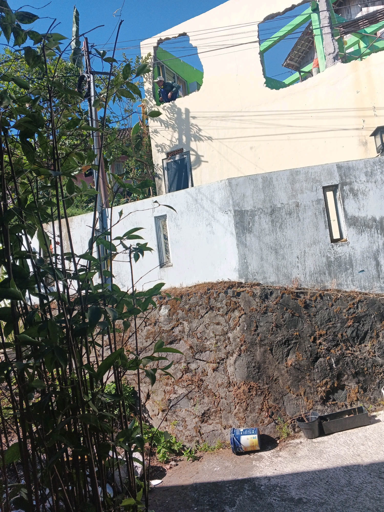 A person wearing a cap and dark clothing stands at a broken window in a partially constructed building in Keji, Indonesia. The building has a yellow facade with a large, irregular opening in the upper story, revealing green structural elements. Below this, a white wall extends across the frame, with a tall, narrow window on the right. In front of the white wall is a rough, dark stone embankment covered in dry vegetation. A cluster of dark green leafy plants grows in the foreground on the left. On the concrete ground in the lower right, there is a blue and white container lying on its side, a black pot with a dry plant, and a black rectangular planter. The sky is clear and blue.