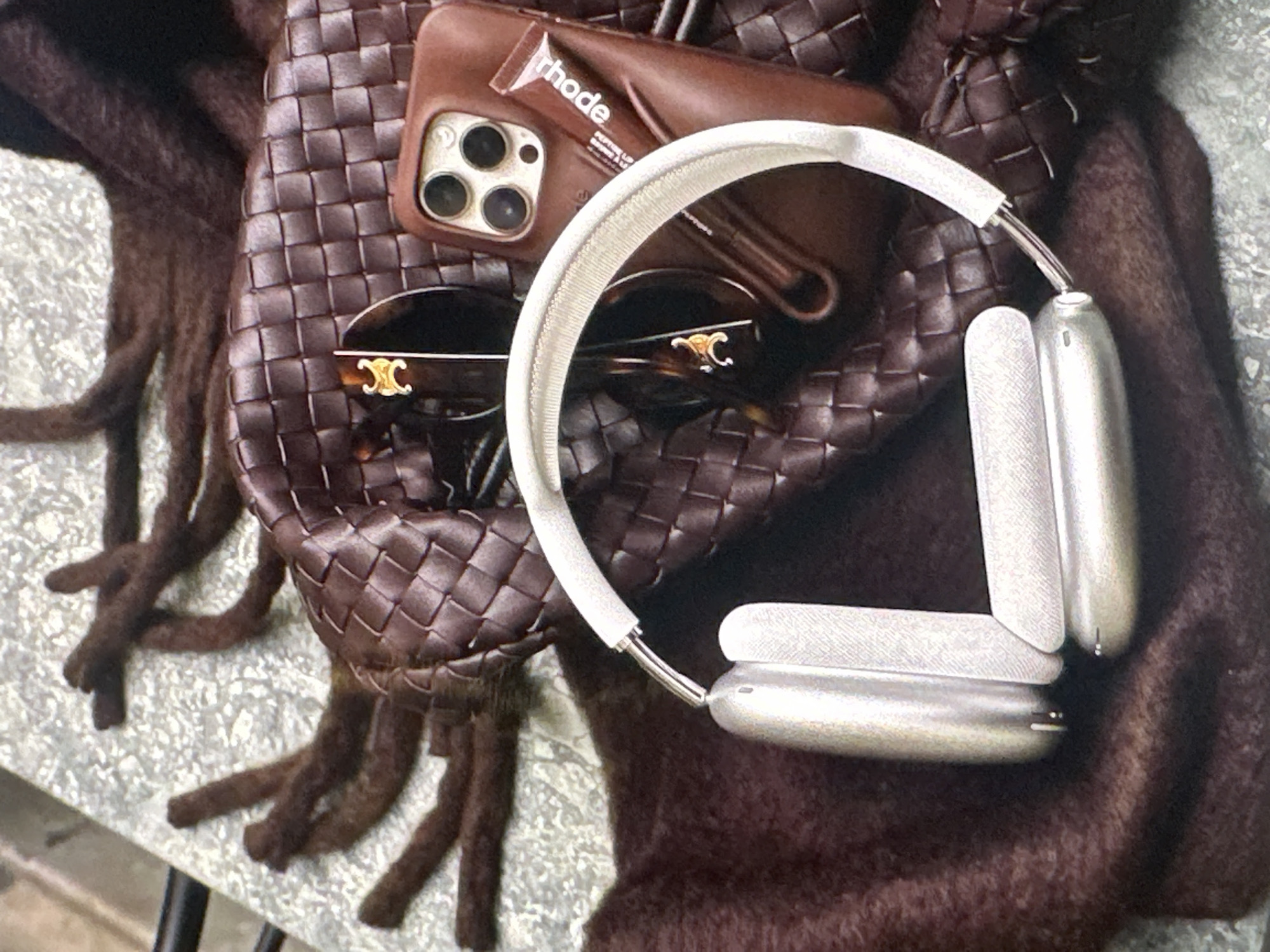 The image presents a collection of luxury items placed on what appears to be a faux fur or fringed textile, possibly brown in color. Dominating the scene are a pair of silver Apple AirPods Max headphones with a white headband. Positioned amongst the headphones and textile is a brown woven handbag or tote. Placed on the bag is a brown phone case housing a smartphone with visible cameras and what looks like a "rhode" branded product. Also on the bag are a pair of Celine sunglasses. There are no people visible in the shot. The setting and location (Yerevan, Armenia) are not evident from the image.