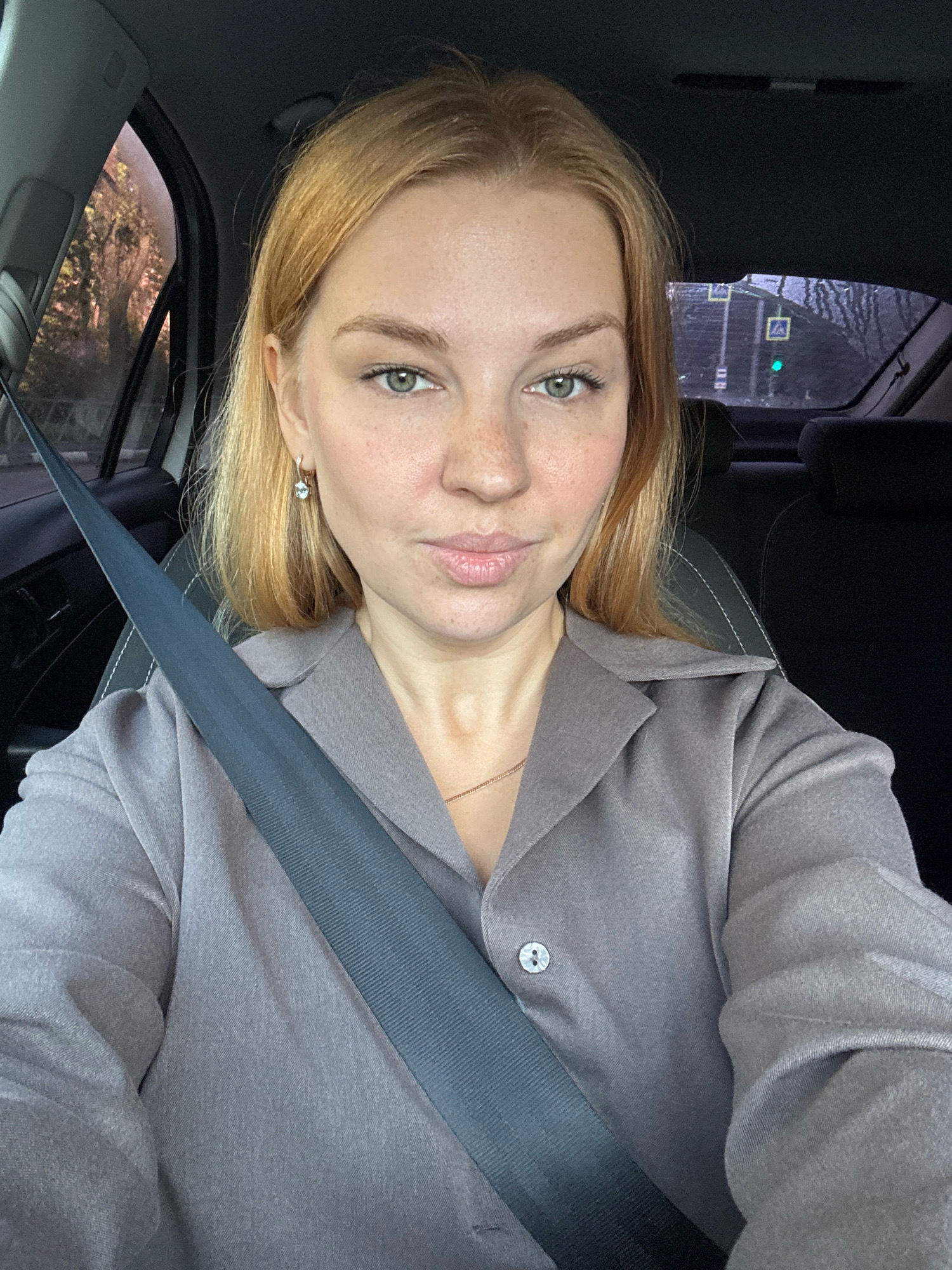 A young woman with blonde hair, freckles, and green eyes is seated in the driver's seat of a car. She is wearing a grey collared shirt with a single button visible, and a grey seatbelt is diagonally across her chest. She is looking directly at the camera with a neutral expression. Outside the car window, trees with autumn foliage are visible, and in the background, a traffic light displays a green signal. The interior of the car, including the headrest of the passenger seat, is also visible. The image appears to have been taken during daylight hours, likely in the city of Rusanovo, Russia.