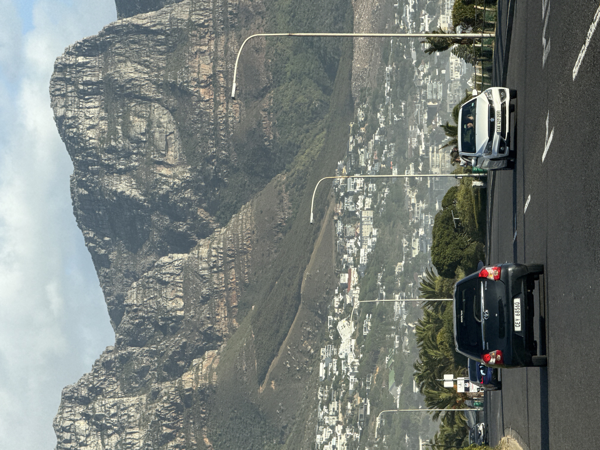 This image shows a road in Cape Town, South Africa, with a mountain range in the background. Two cars are visible: a white car and a black car, each traveling on the road. The mountain face has distinct layers, indicating its geological formation. On the right side of the road, there's a densely populated urban area with white buildings and greenery. Tall street lamps are situated along the roadside, and the weather appears to be sunny.