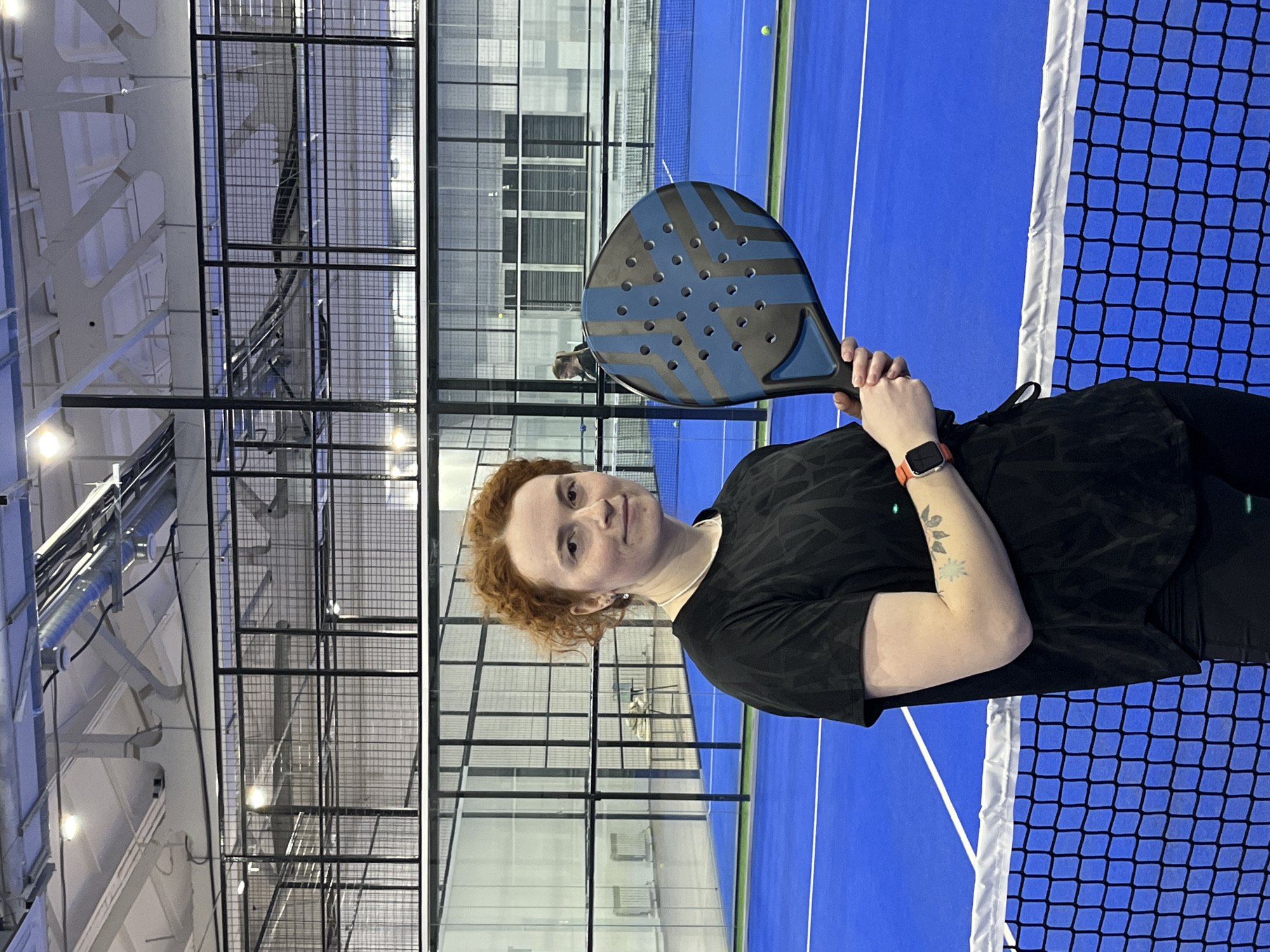 A woman with curly red hair is standing indoors on a blue tennis court in what appears to be a padel court. She's holding a padel racket with blue and black details. She's wearing a black t-shirt and a red smartwatch. The court has black wire mesh enclosures. The location is likely in a city with modern architecture, possibly Chaikovsky, Russia.