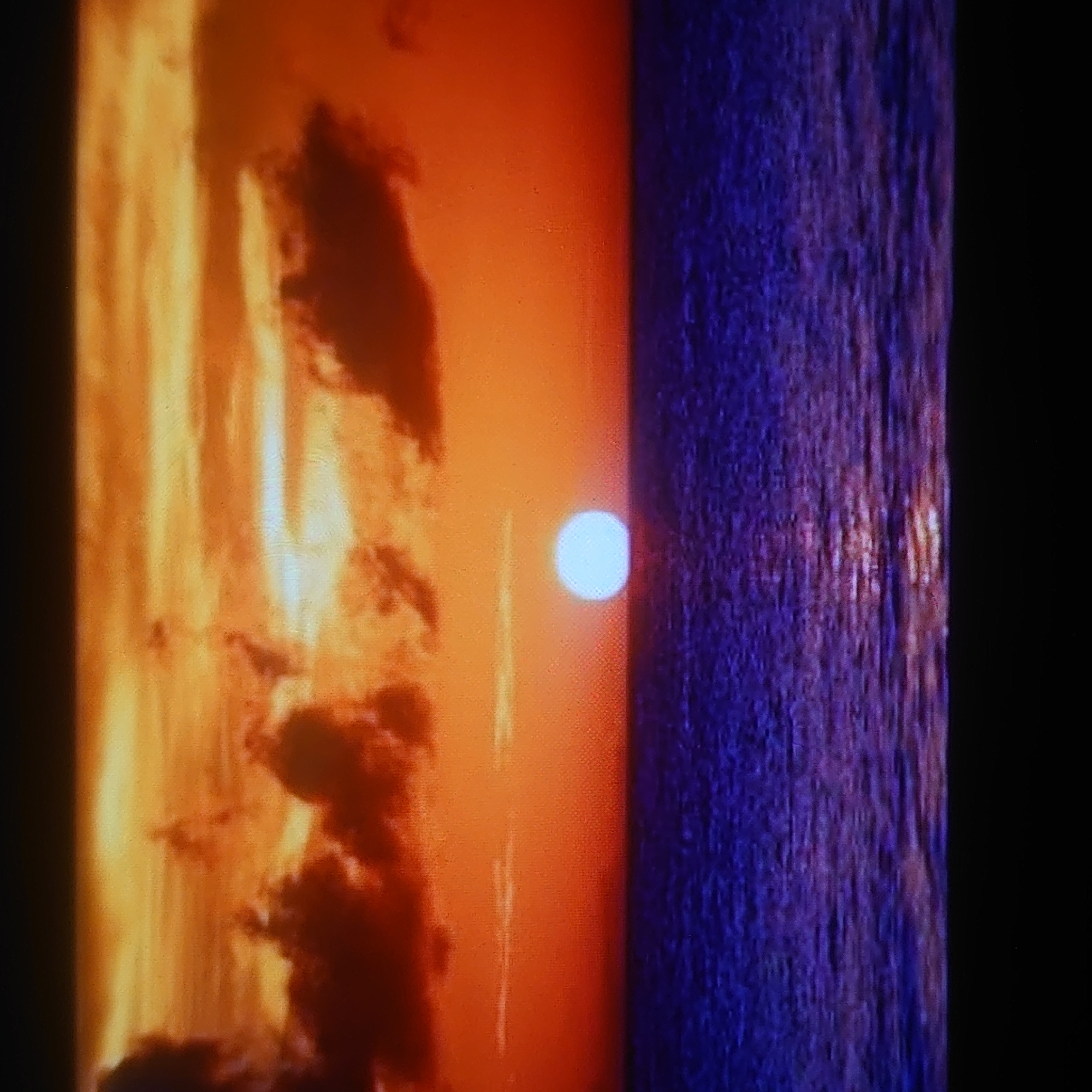 The image shows a sunset over a body of water. The sky is a vibrant orange and yellow, with dark clouds scattered throughout. The sun is a bright white orb resting on the horizon. The water is a deep blue, with reflections of the sky and sun rippling across its surface. There are no people or objects visible in the image. The location is not specified in the image, and it is impossible to determine if it is in Belorechensk, Russia.